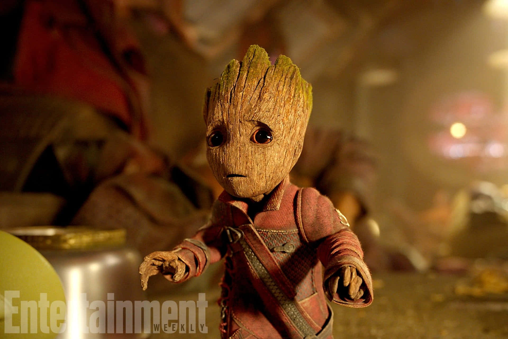 Guardians Of The Galaxy 2 桌布 壁紙 — 一個細小、像樹一樣的生物，有大眼睛和紅色衣服，站在一個光線昏暗、雜亂的房間裡。