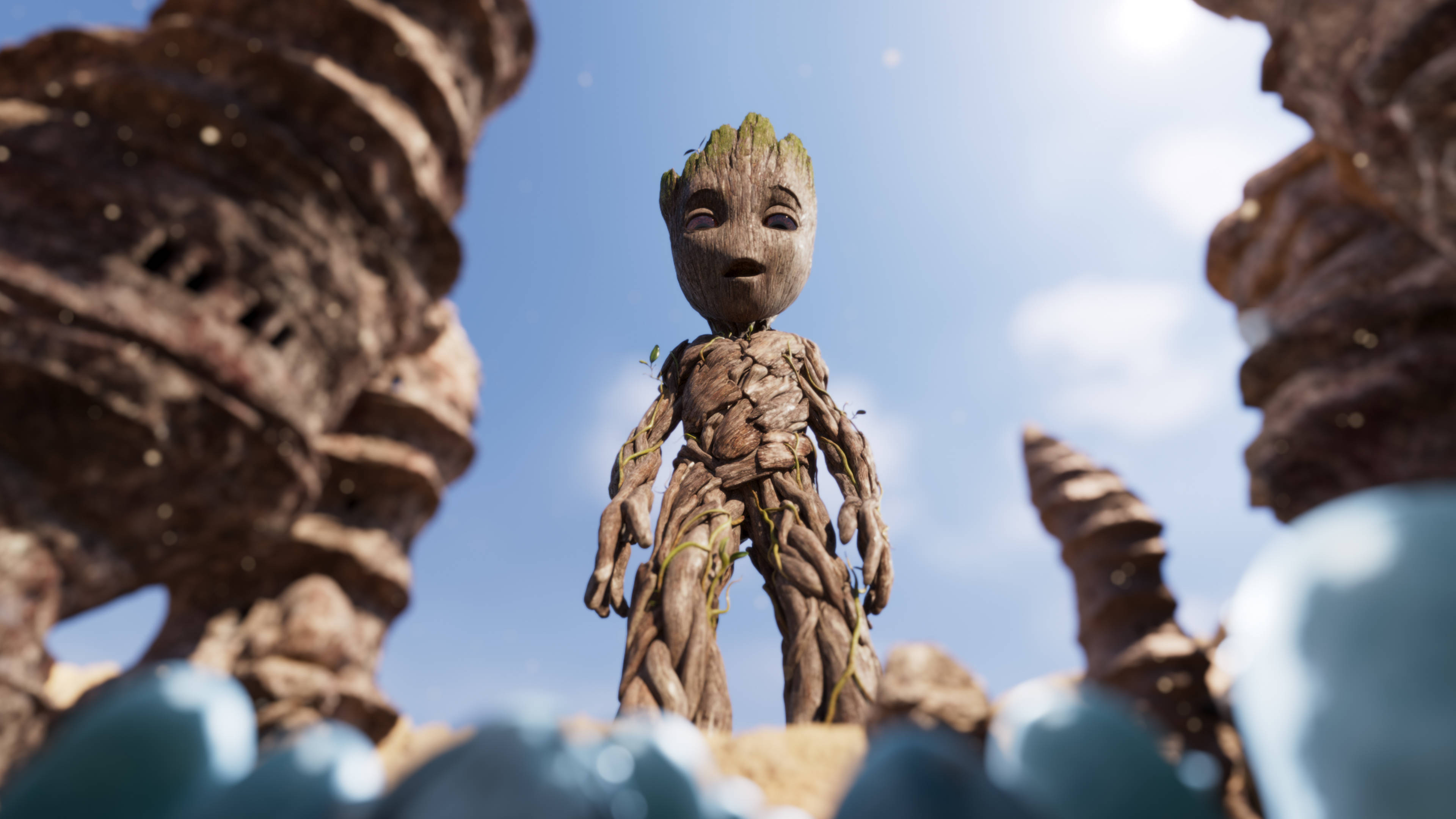 Baby Groot 桌布 壁紙 — 一個細小的樹狀生物站在巨大岩石形成物之間，在有散雲的明亮藍天下。
