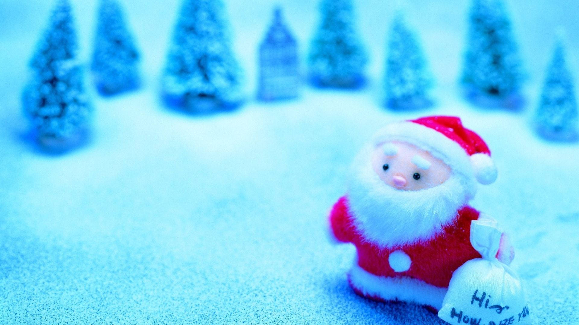 Kawaii Christmas 桌布 壁紙 — 一個細小的毛絨聖誕老人拿著禮物袋，站在一個有微型樹木的雪藍色風景中。