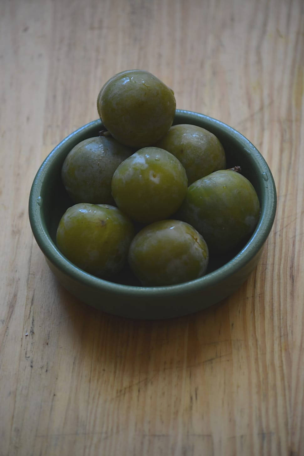 Olive 桌布 壁紙 — 一個細小的綠色陶瓷碗，裝滿了飽滿的綠色李子，放在溫暖的木質表面上。