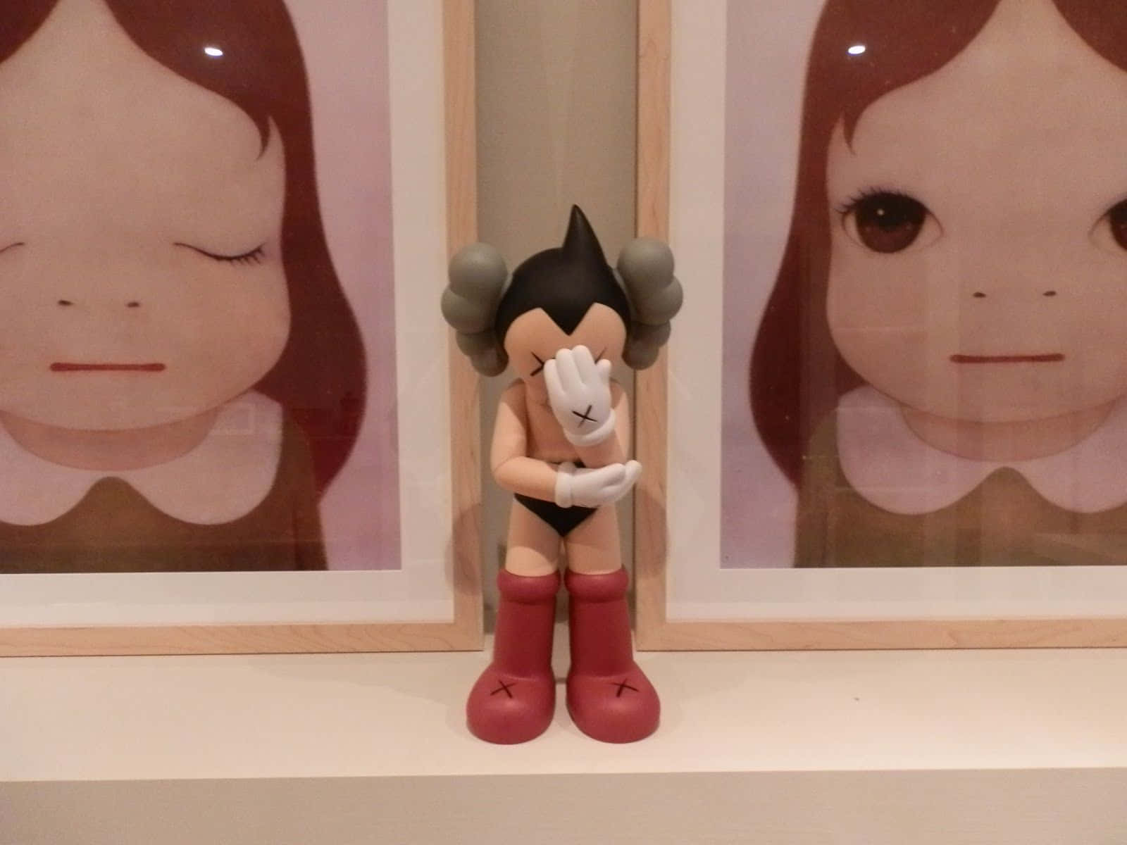 Kaws Astro Boy 桌布 壁紙 — 一個細小的黑紅色KAWS公仔站在一個架子上，位於兩個棕色頭髮女孩的裱框肖像之間。