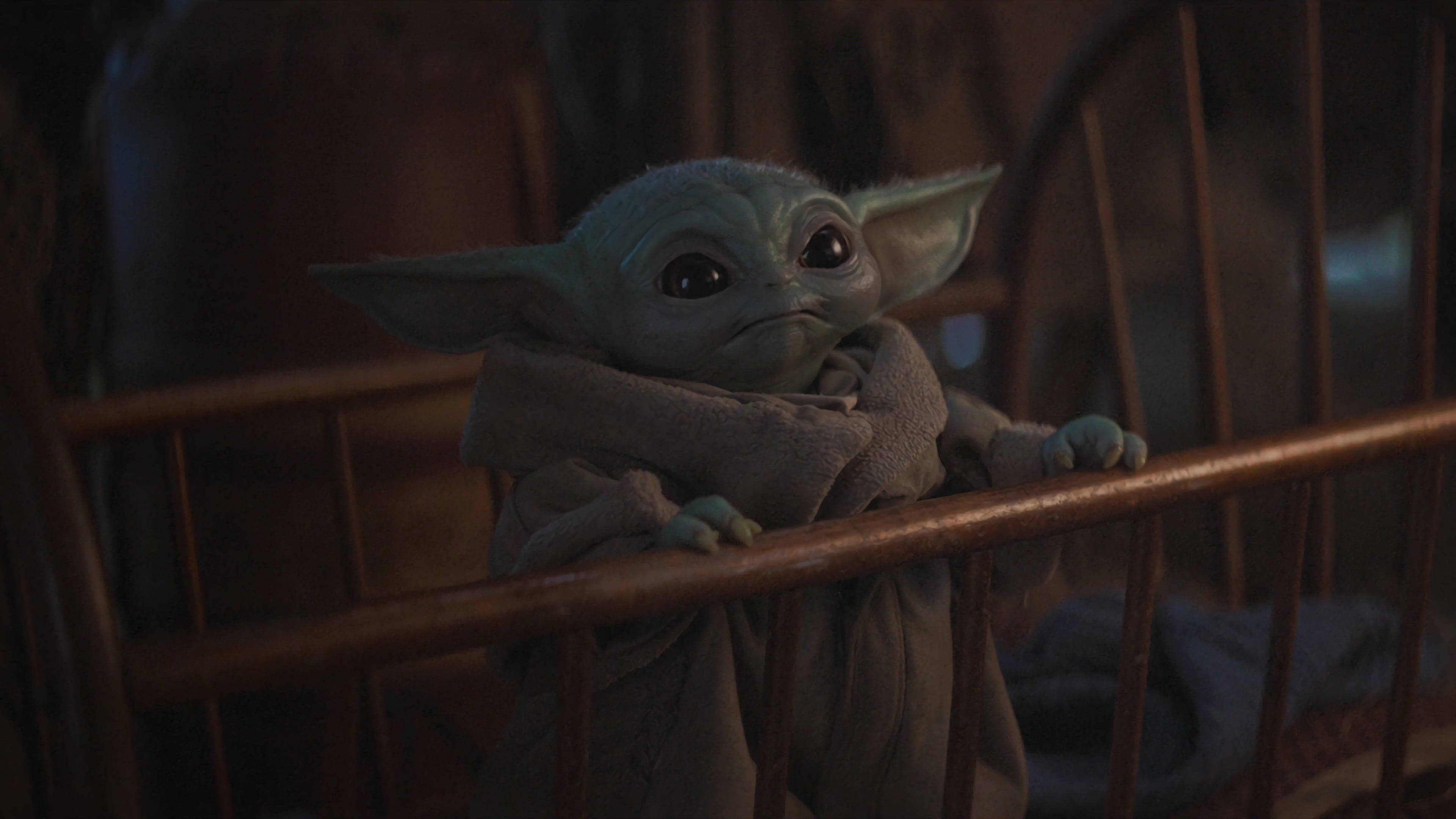 Mandalorian And Baby Yoda 桌布 壁紙 — 一個細小綠色外星人，有大耳朵和大眼睛，在昏暗燈光下從木製嬰兒床欄杆上方窺視。