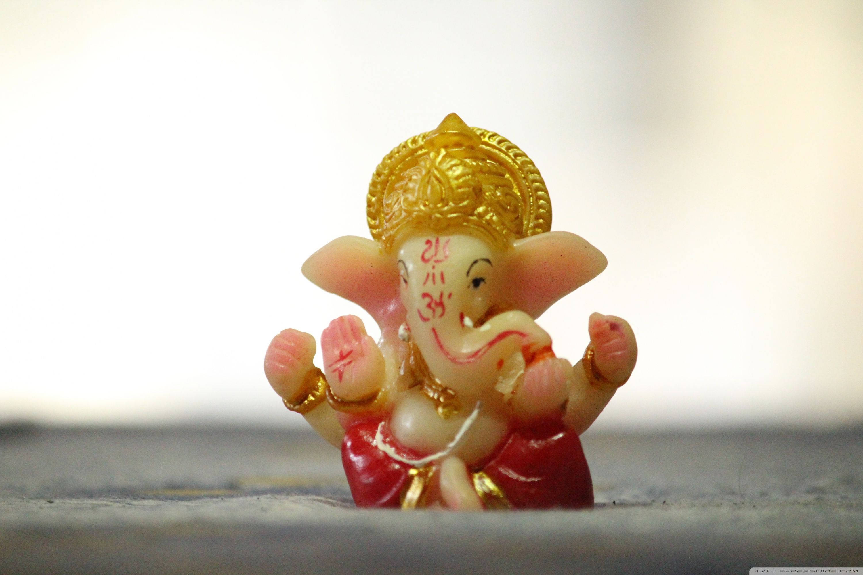 cute ganesha 桌布 壁紙 — 一個細小色彩繽紛的象神像，戴著金色皇冠和紅色衣服，微笑著坐在柔和的模糊背景前。
