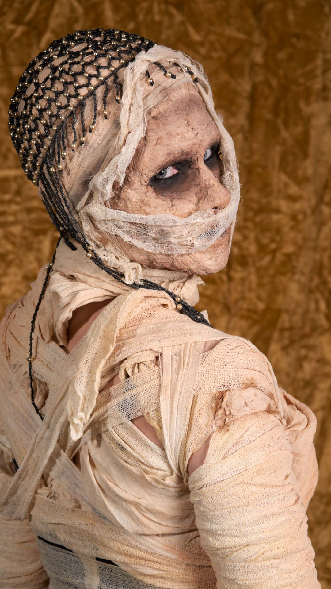 Mummy Costumes 桌布 壁紙 — 一個細節令人毛骨悚然的木乃伊，纏著繃帶的臉龐和華麗的頭飾，在紋理豐富的金色背景下強烈凝視。