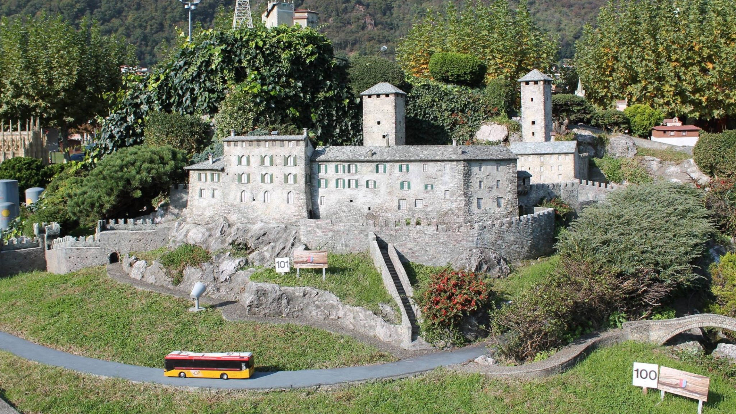 Switzerland 桌布 壁紙 — 一個細節豐富的微型石城堡，擁有塔樓和城牆，坐落在茂密的綠植中，蜿蜒的小路上有一輛小型巴士。