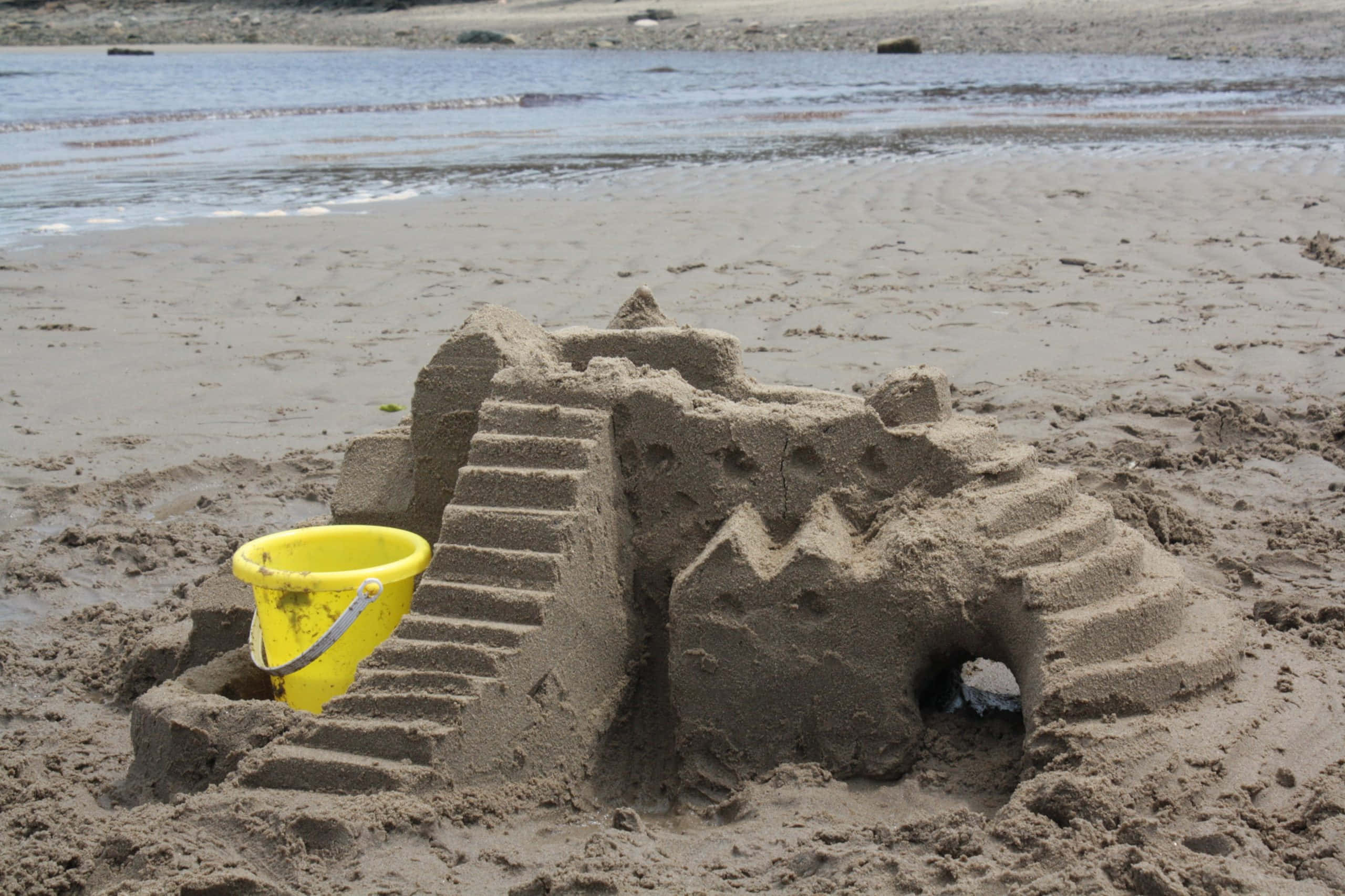 Sandcastles 桌布 壁紙 — 一個細節豐富的沙堡，帶有樓梯和塔樓，矗立在濕漉漉的海灘上，旁邊有一個亮黃色的水桶。