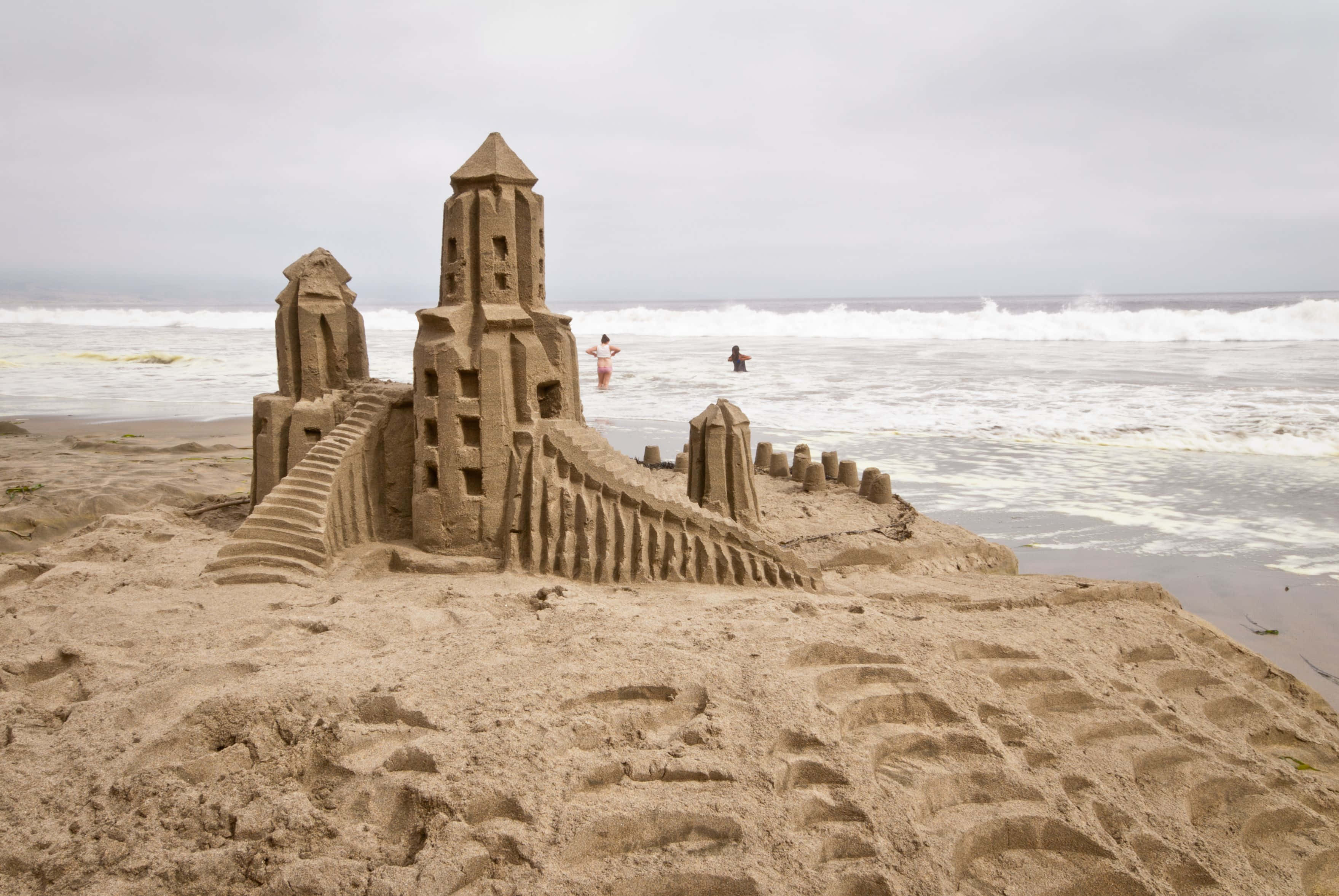 Sandcastles 桌布 壁紙 — 一個細節豐富的沙堡，有塔樓和樓梯，矗立在沙灘上，兩人在多雲的天空下在海中游泳。