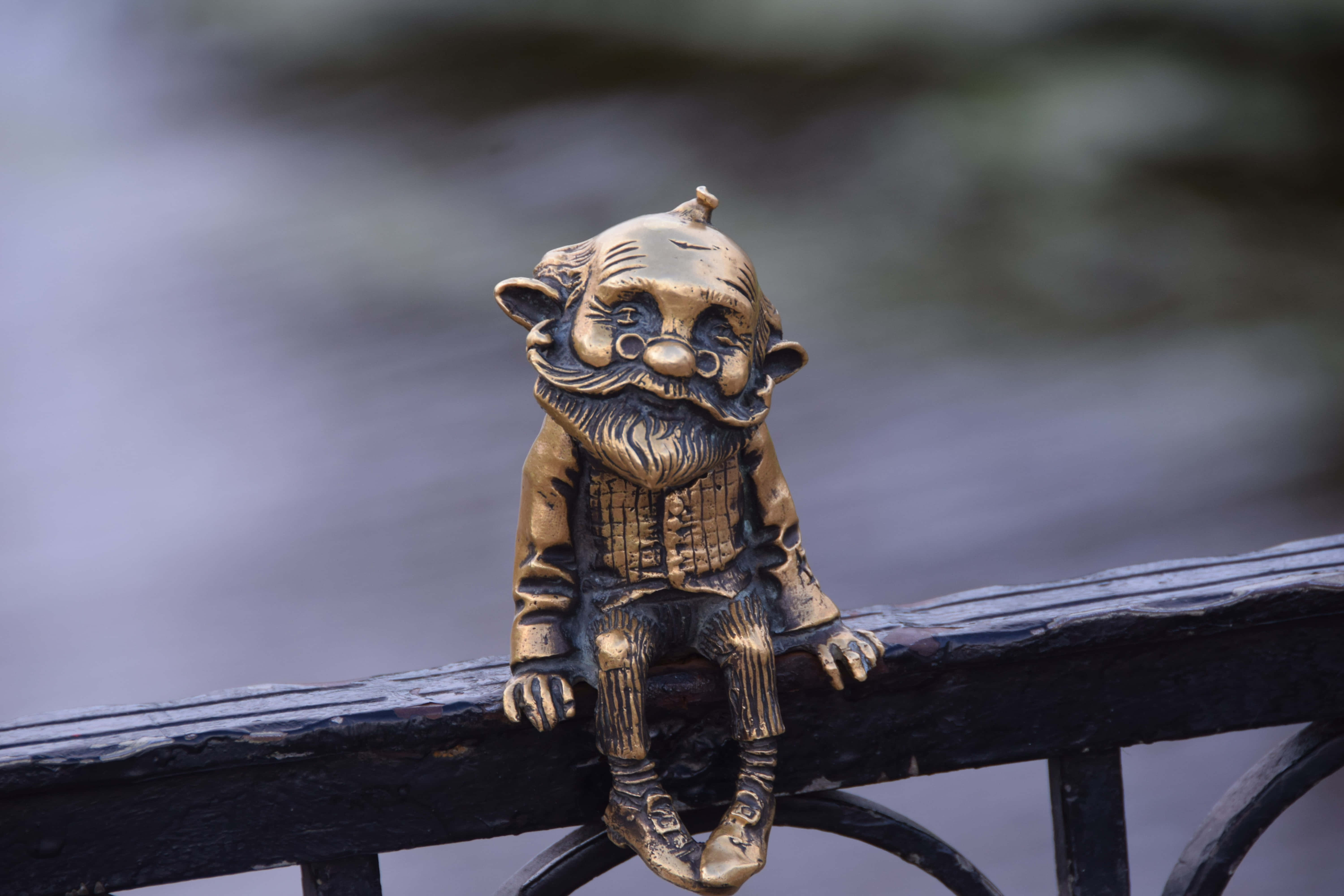 Bronze 桌布 壁紙 — 一個細節豐富的黃銅花園地精，長鬍鬚戴眼鏡，坐在俯瞰水面的黑色金屬欄杆上。