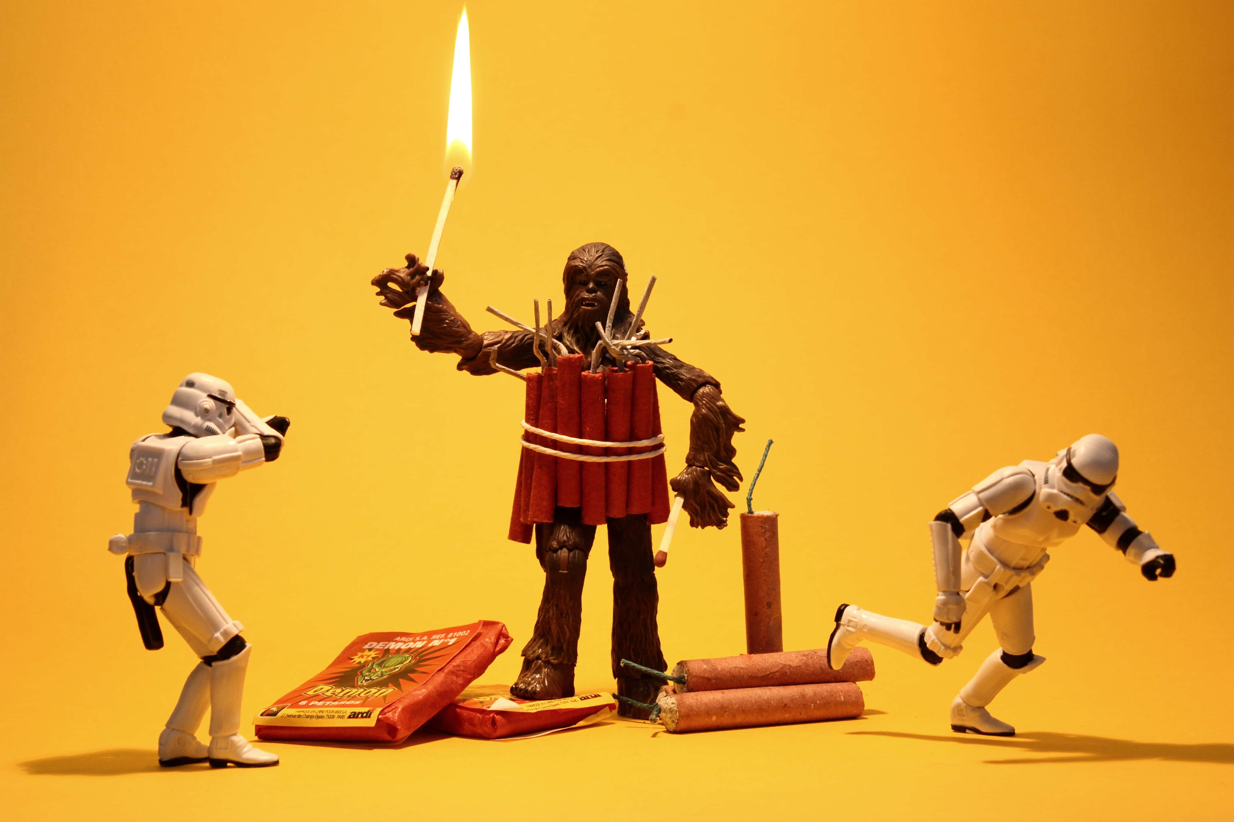 Wookiee 桌布 壁紙 — 一個綁著紅色爆竹的丘巴卡動作人偶手持點燃的引信，兩個風暴兵人偶在純黃色背景前戲劇性地反應。