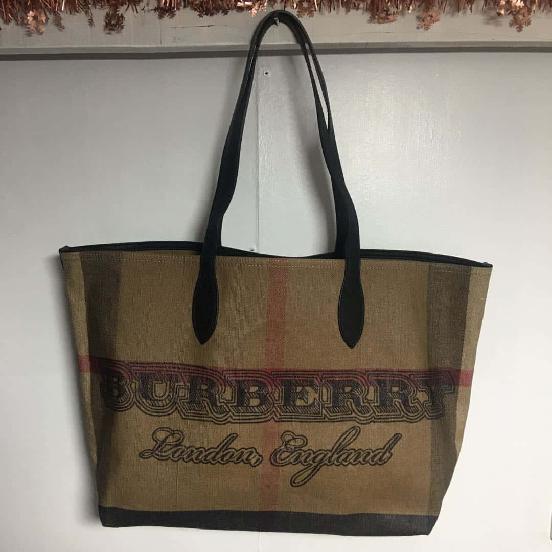 Burberry 桌布 壁紙 — 一個經典格紋的Burberry托特包掛在純白牆上，下方有銅色閃片裝飾。