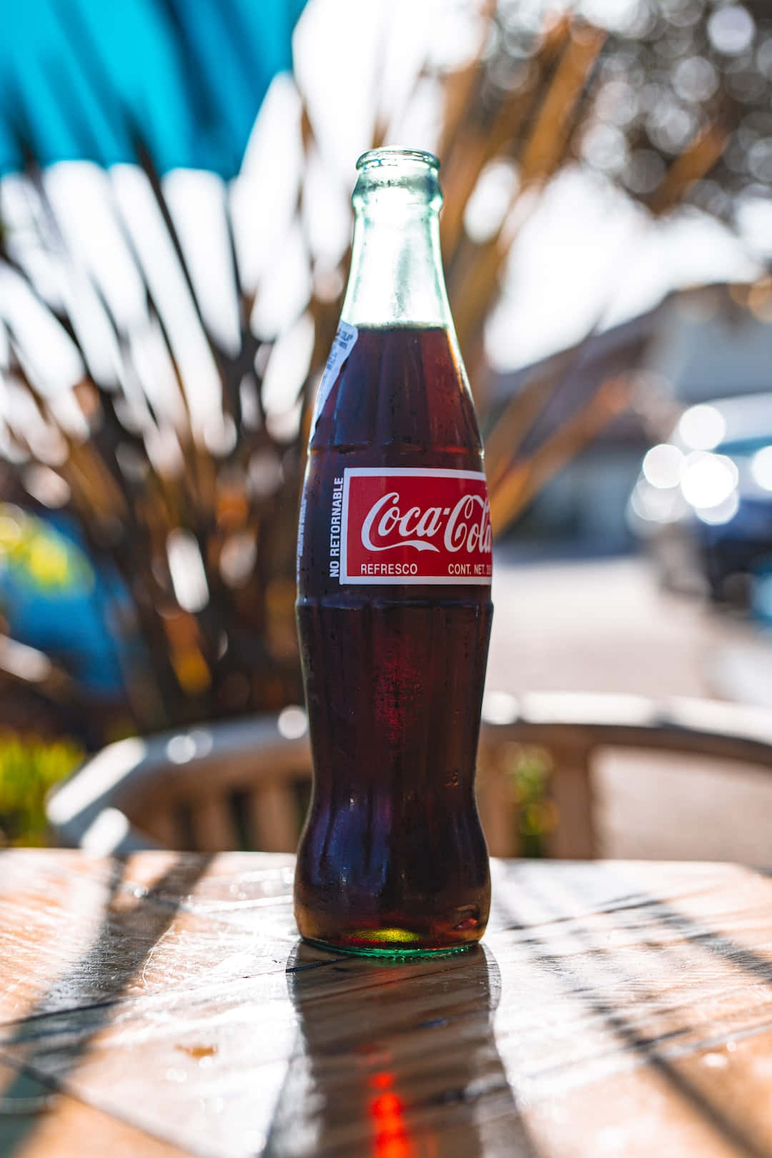 coca cola 桌布 壁紙 — 一個經典的可口可樂瓶放在陽光照射的木桌上，背景有棕櫚葉和藍色雨傘模糊不清。
