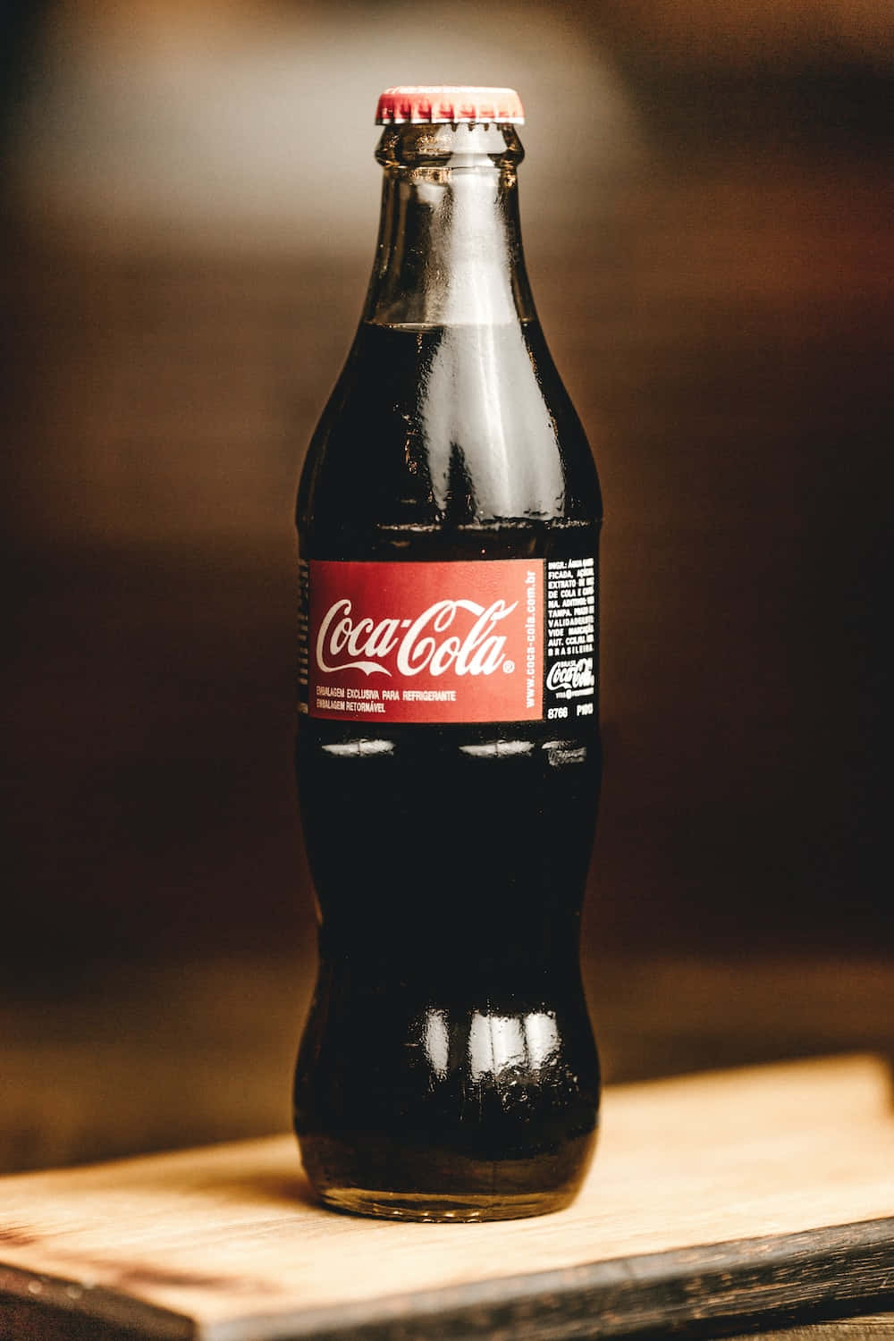 coca cola 桌布 壁紙 — 一個經典的玻璃可口可樂瓶，帶有紅色標籤，放在木質表面上，背景溫暖模糊。