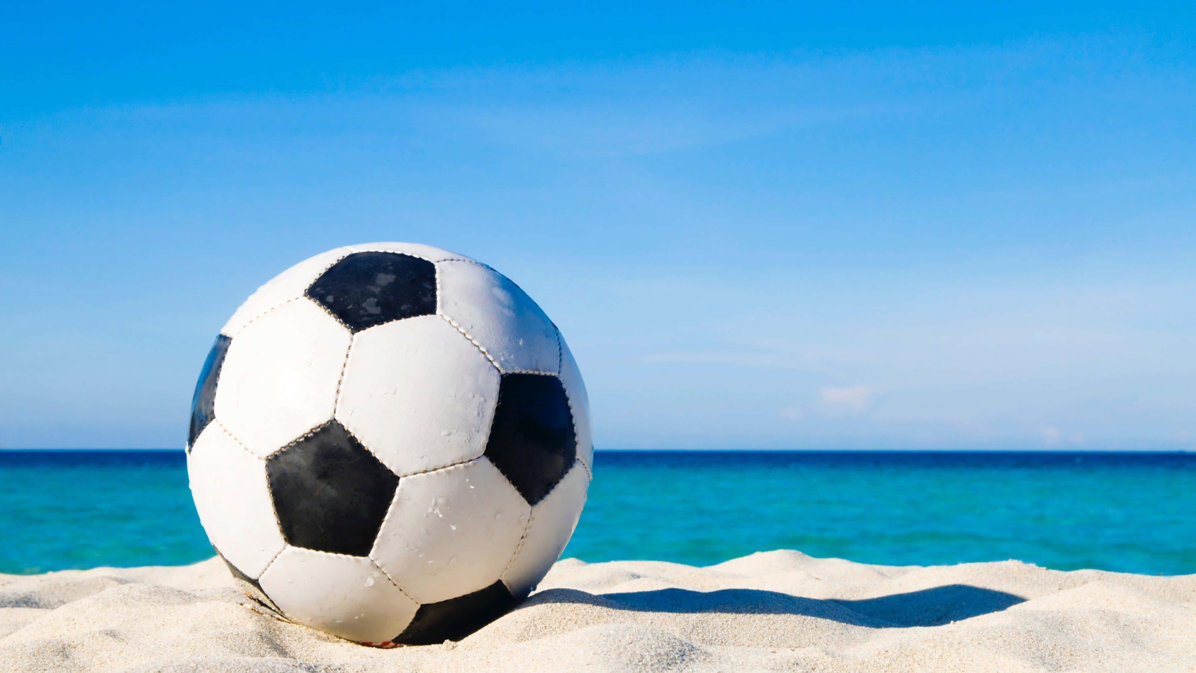 Soccer Ball 桌布 壁紙 — 一個經典的黑白足球擱在陽光照耀的沙灘上，背後是碧綠色的海洋和清澈的藍天。