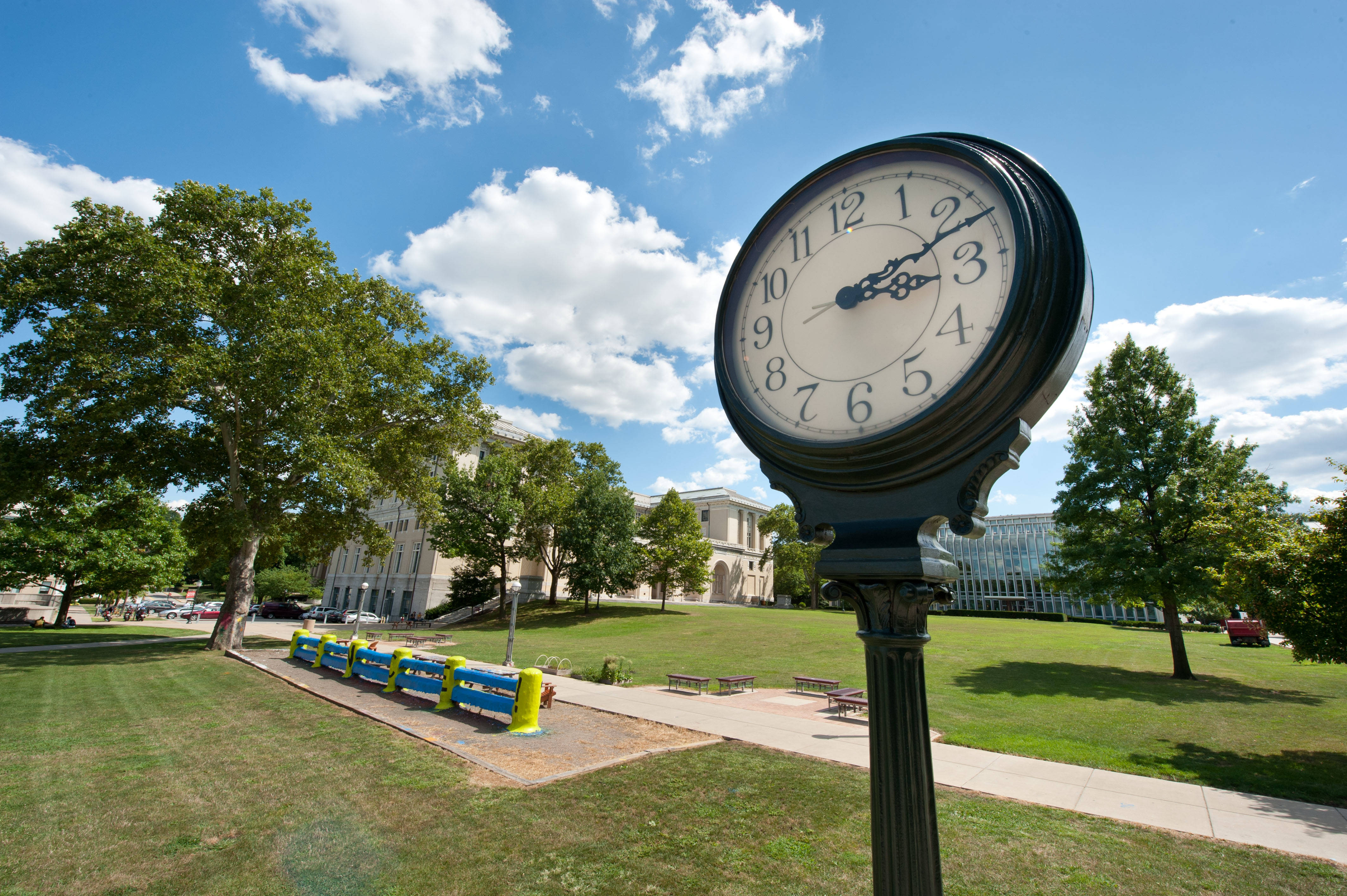 Clock 桌布 壁紙 — 一個經典的黑色街頭時鐘矗立在陽光明媚的公園中，周圍有綠草、樹木和一座宏偉的建築，藍天白雲。