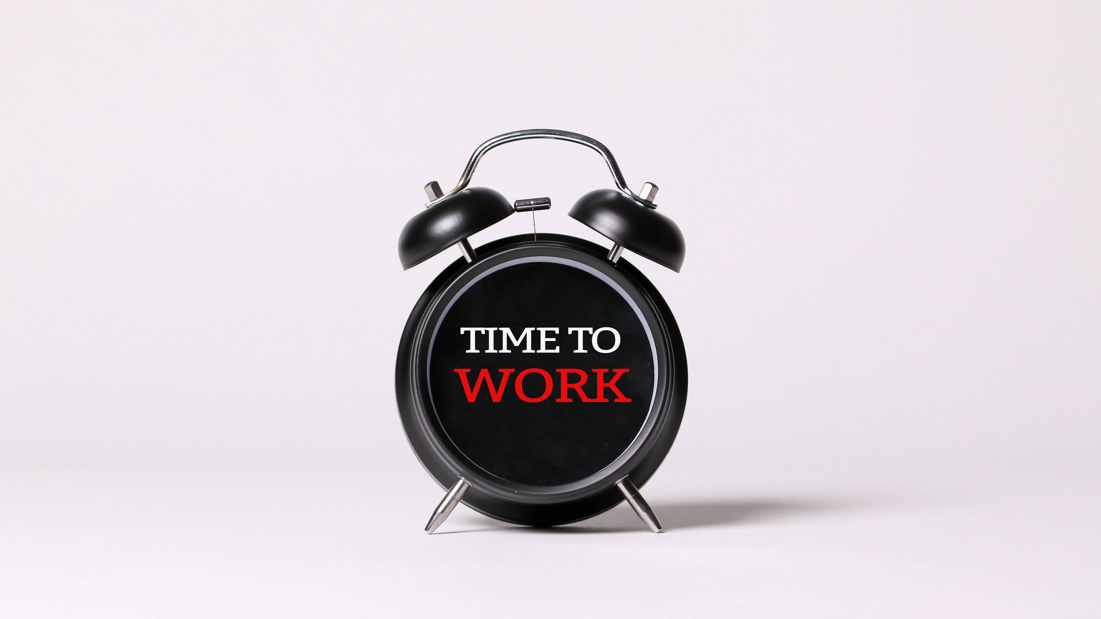 Alarm 桌布 壁紙 — 一個經典的黑色鬧鐘，帶有白色和紅色的「TIME TO WORK」文字，立在乾淨的白色背景前。