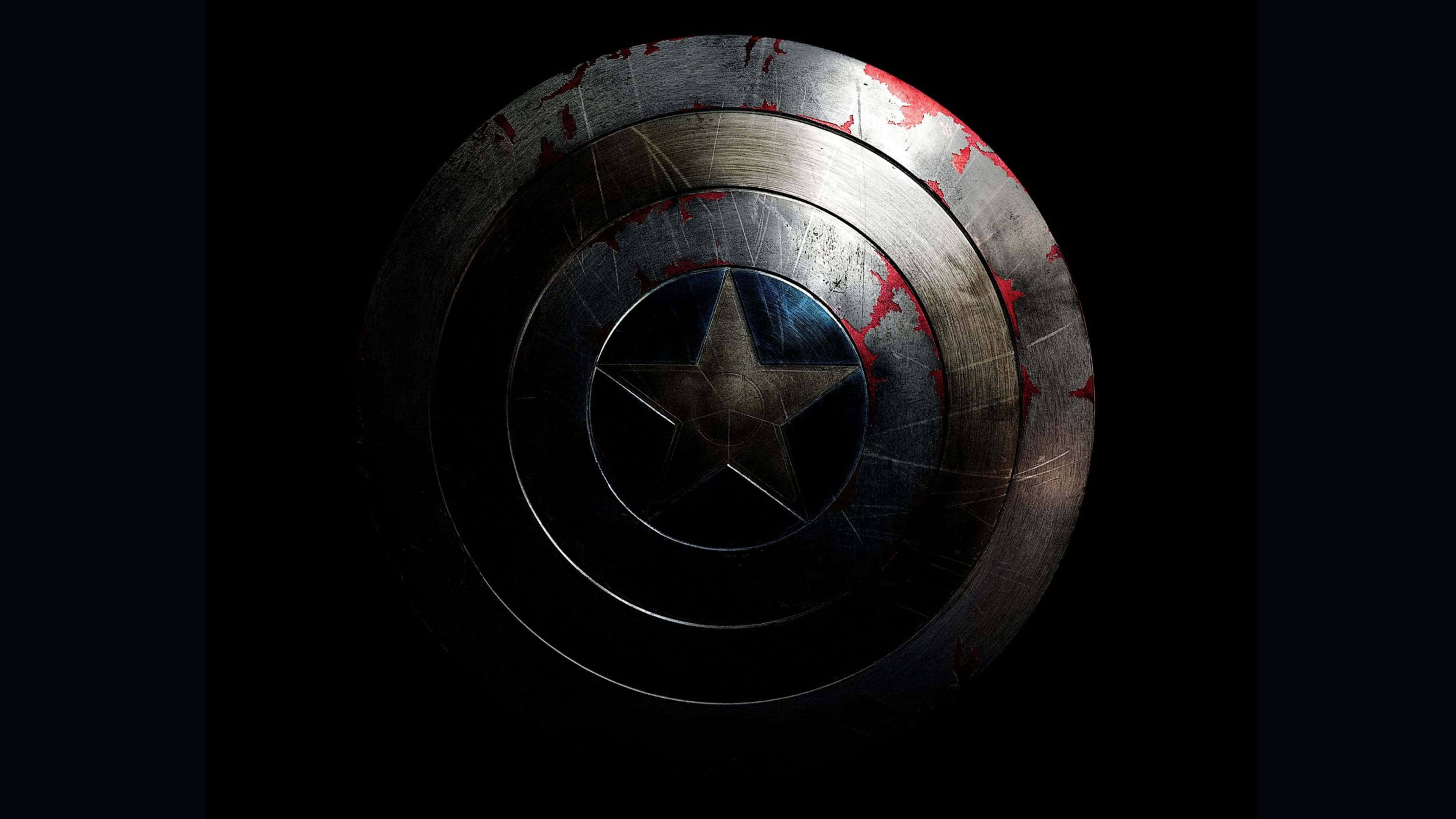 4K Captain America 桌布 壁紙 — 一個經過戰鬥磨損的美國隊長盾牌，帶有紅色飛濺和刮痕，在黑色背景的戲劇性燈光下發光。