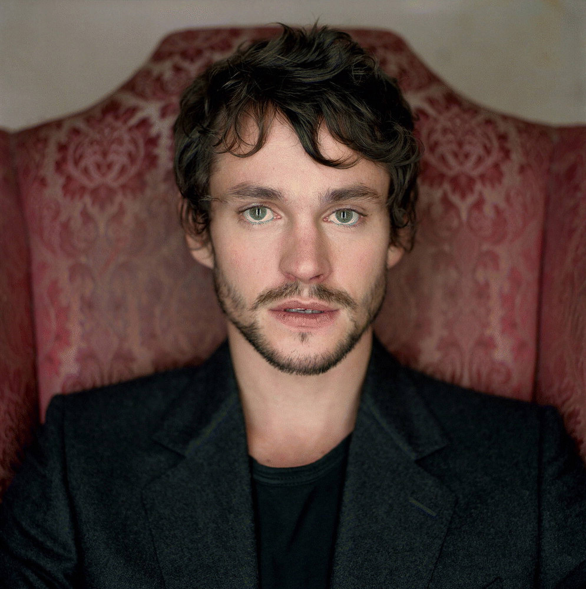 Hugh Dancy 桌布 壁紙 — 一個綠眼睛黑頭髮的男人坐在紅色圖案扶手椅上，穿著深色西裝。
