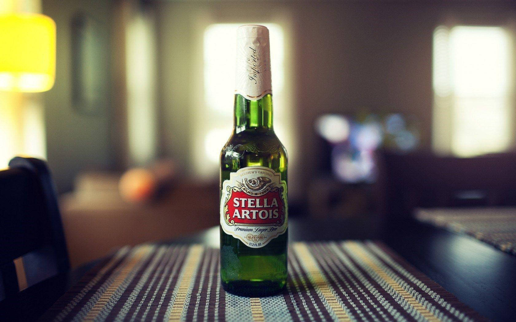 飲料 桌布 壁紙 — 一個綠色 Stella Artois 啤酒瓶放在條紋桌布中央，處於溫暖照明、柔和模糊的客廳中。