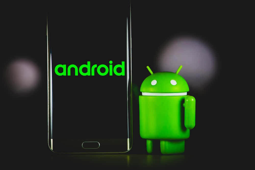 安卓 桌布 壁紙 — 一個綠色Android吉祥物公仔站在一部顯示Android標誌的智慧型手機旁邊，背景為深色模糊。