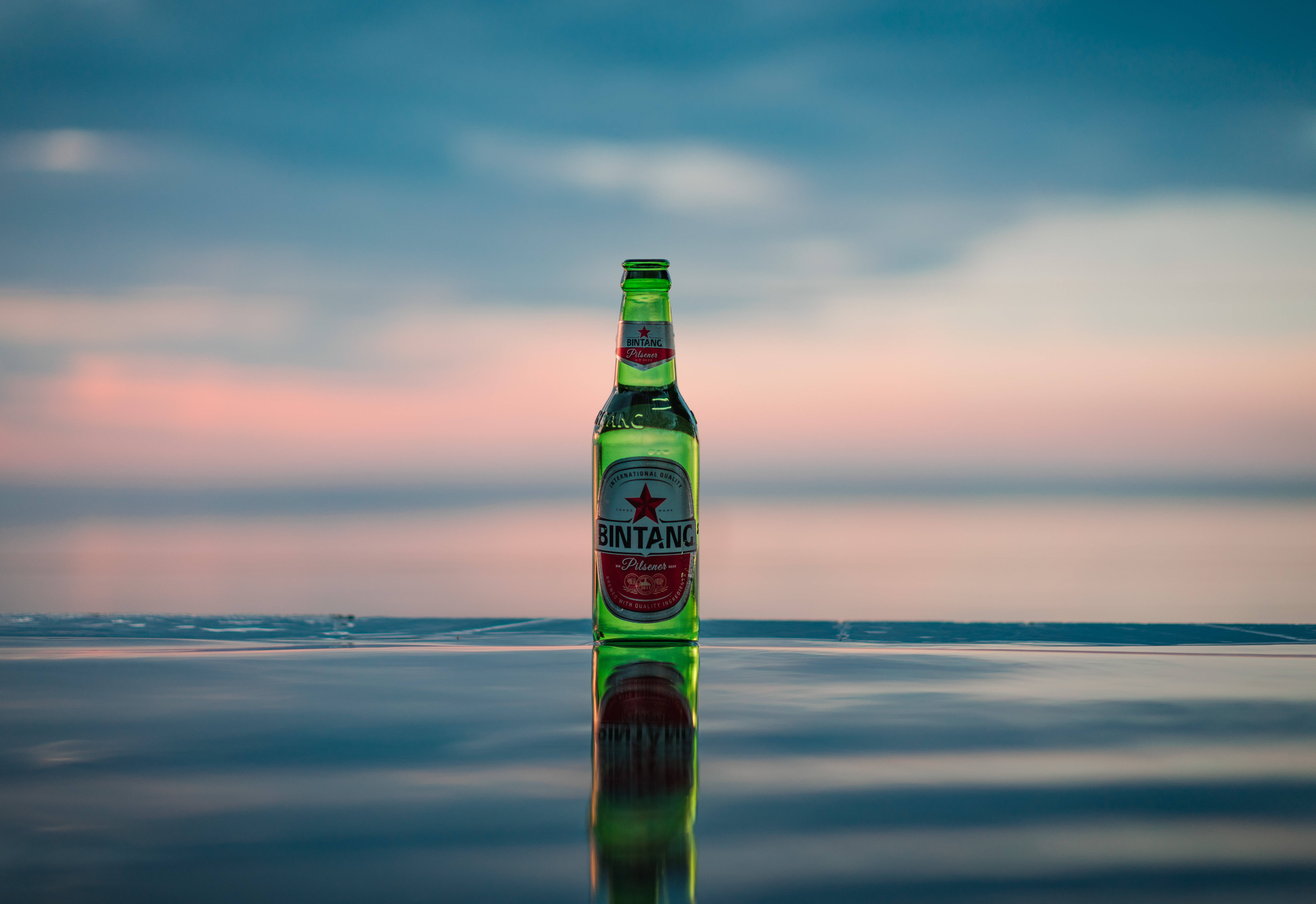 Beer 桌布 壁紙 — 一個綠色Bintang啤酒瓶直立於平靜的水面上，水面反射出柔和的粉紅色和藍色的黃昏天空。