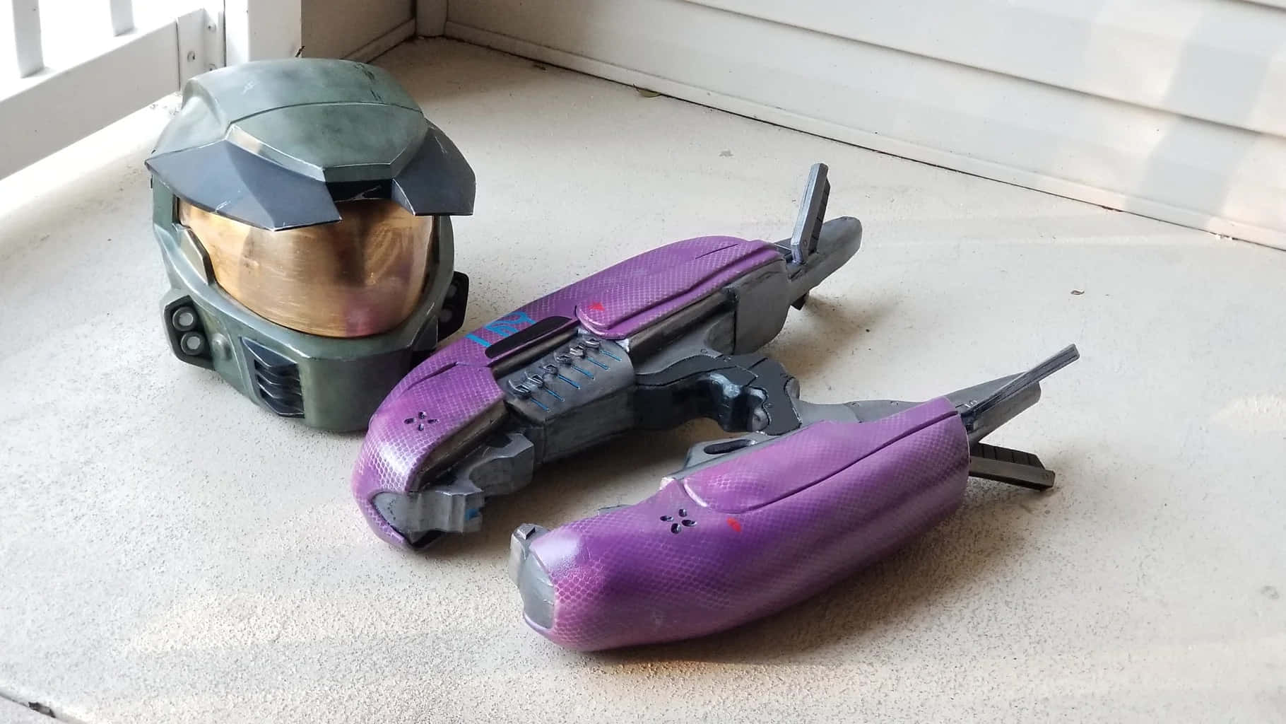 Plasma Rifle 桌布 壁紙 — 一個綠色Halo頭盔和兩把紫色能量武器放在窗邊淺灰色混凝土的地板上。