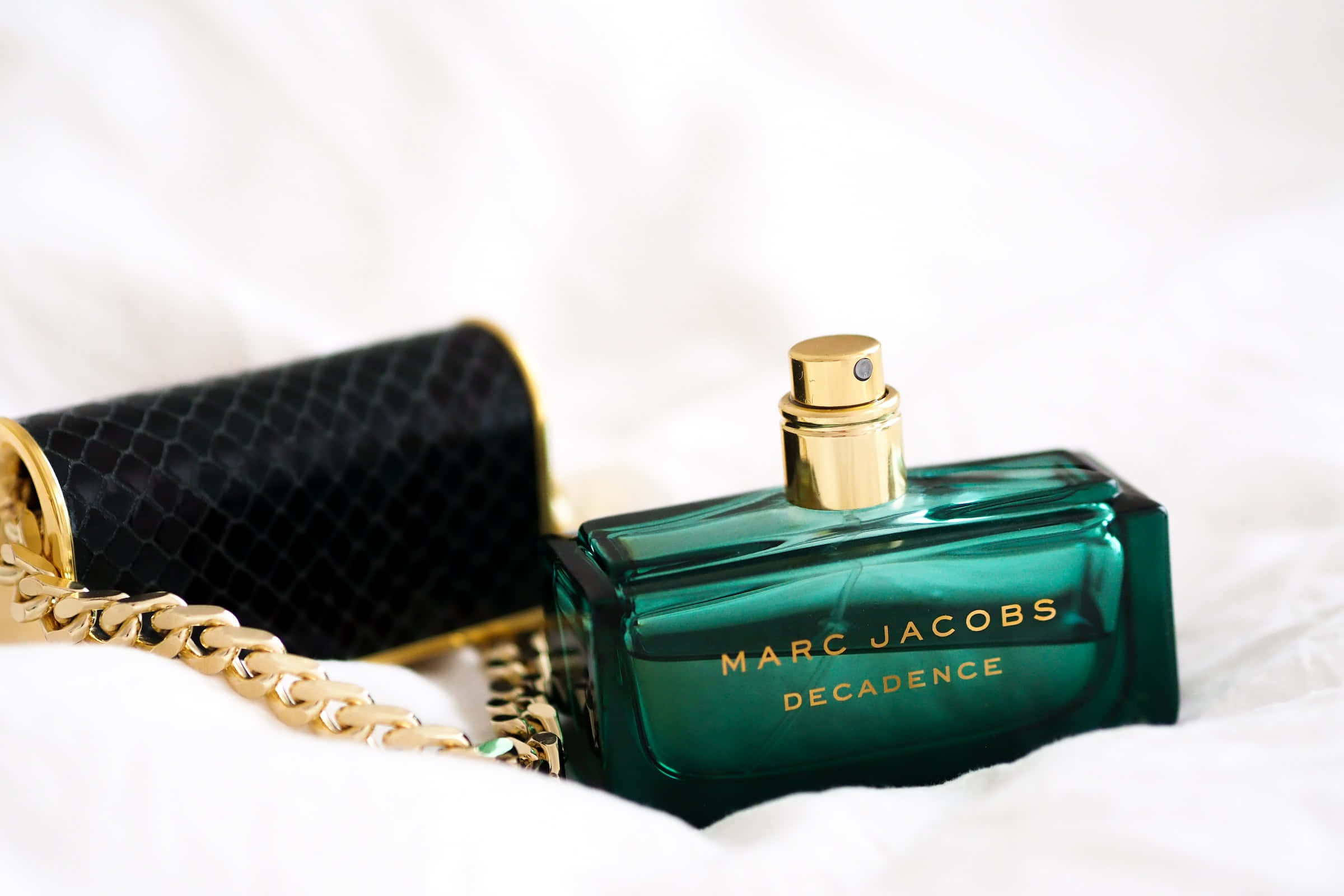 Aesthetic Green 桌布 壁紙 — 一個綠色Marc Jacobs Decadence香水瓶，金色瓶蓋，旁邊放著一個黑色蛇皮手拿包，置於白色布料上。