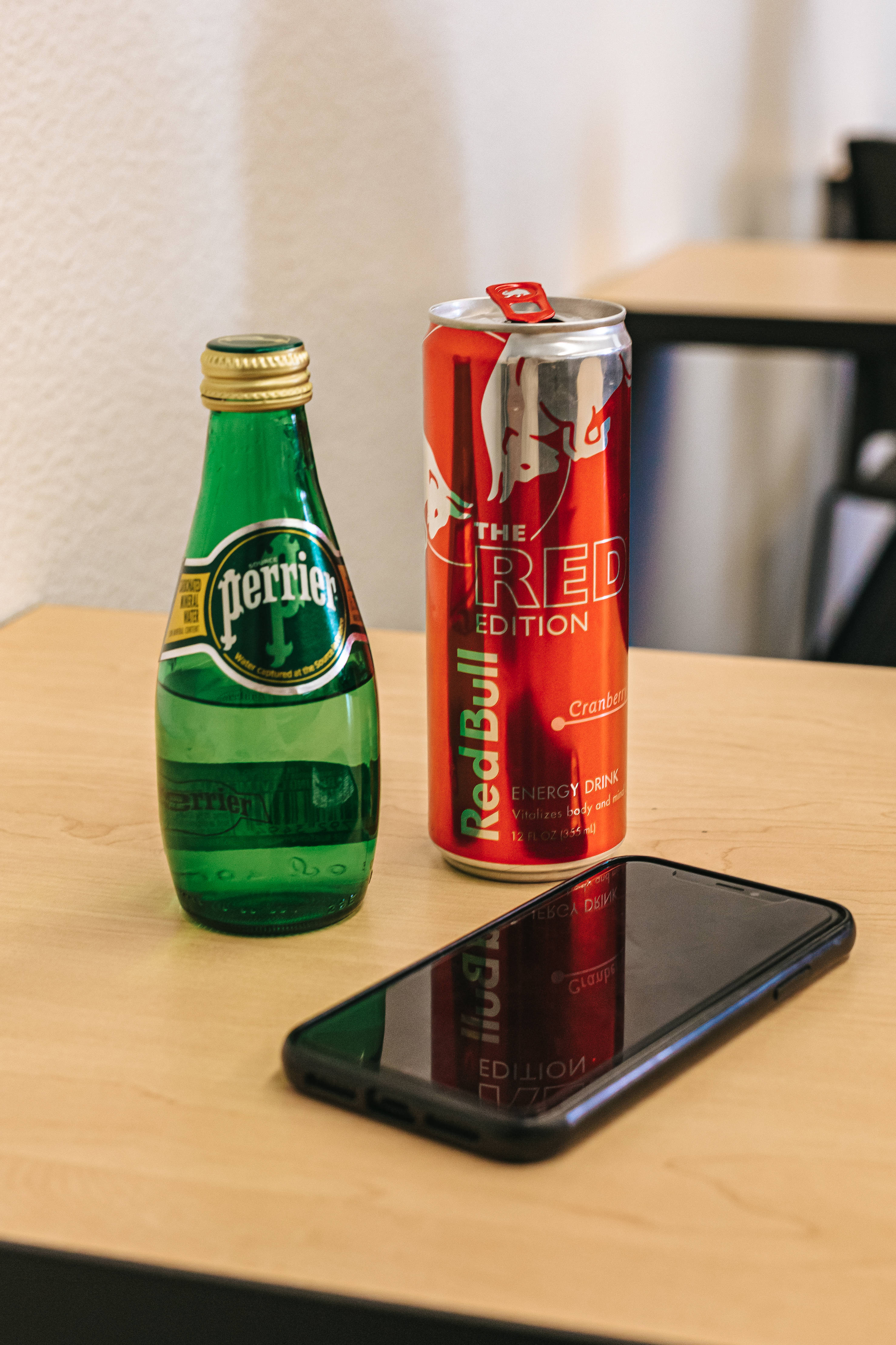 bull 桌布 壁紙 — 一個綠色Perrier瓶和一個紅色Red Bull罐放在淺色木桌上，旁邊是一部黑色手機。