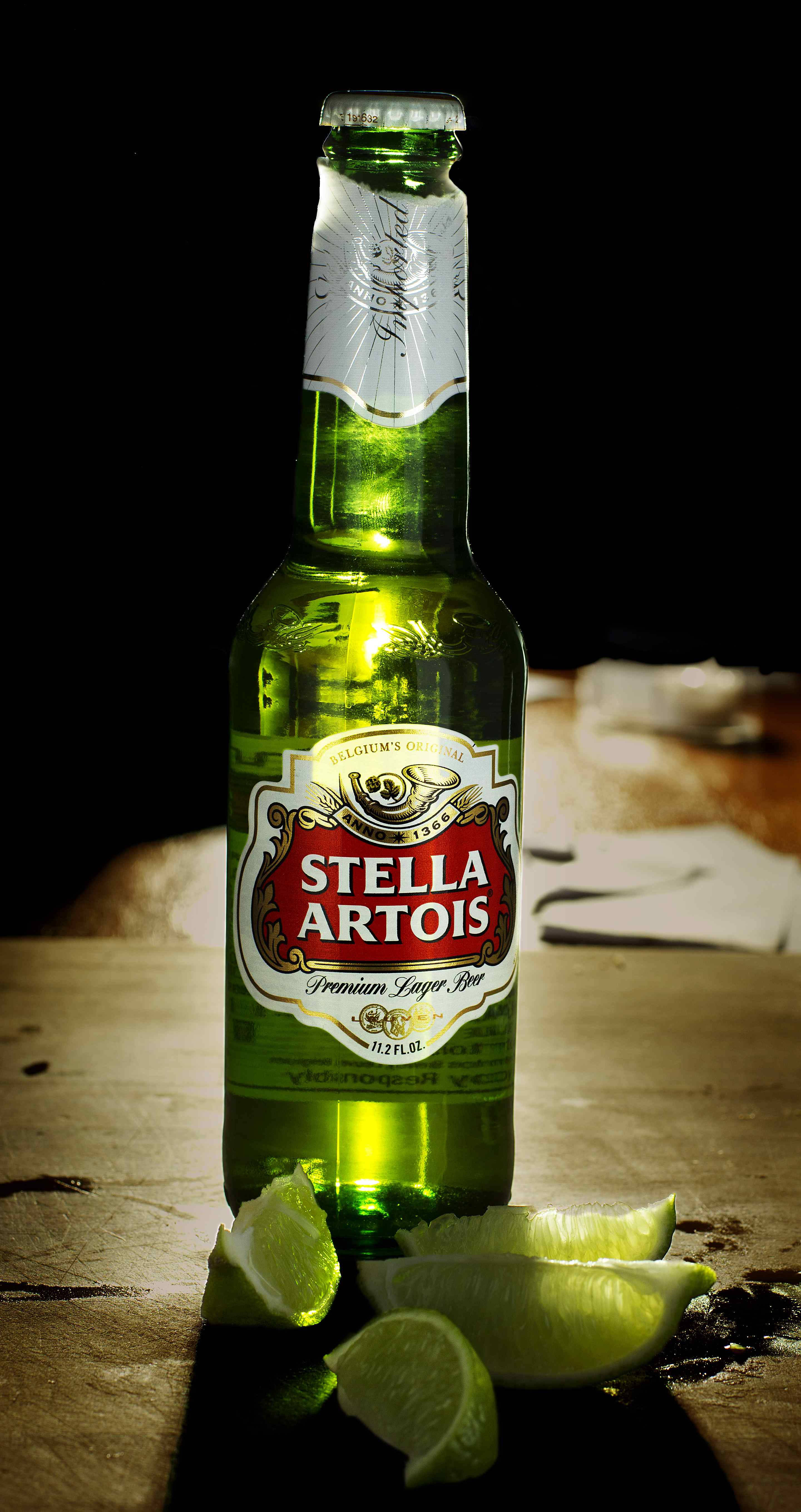 Drink 桌布 壁紙 — 一個綠色Stella Artois啤酒瓶配以萊姆角，放在木桌上，黑色背景下的戲劇性聚光燈。