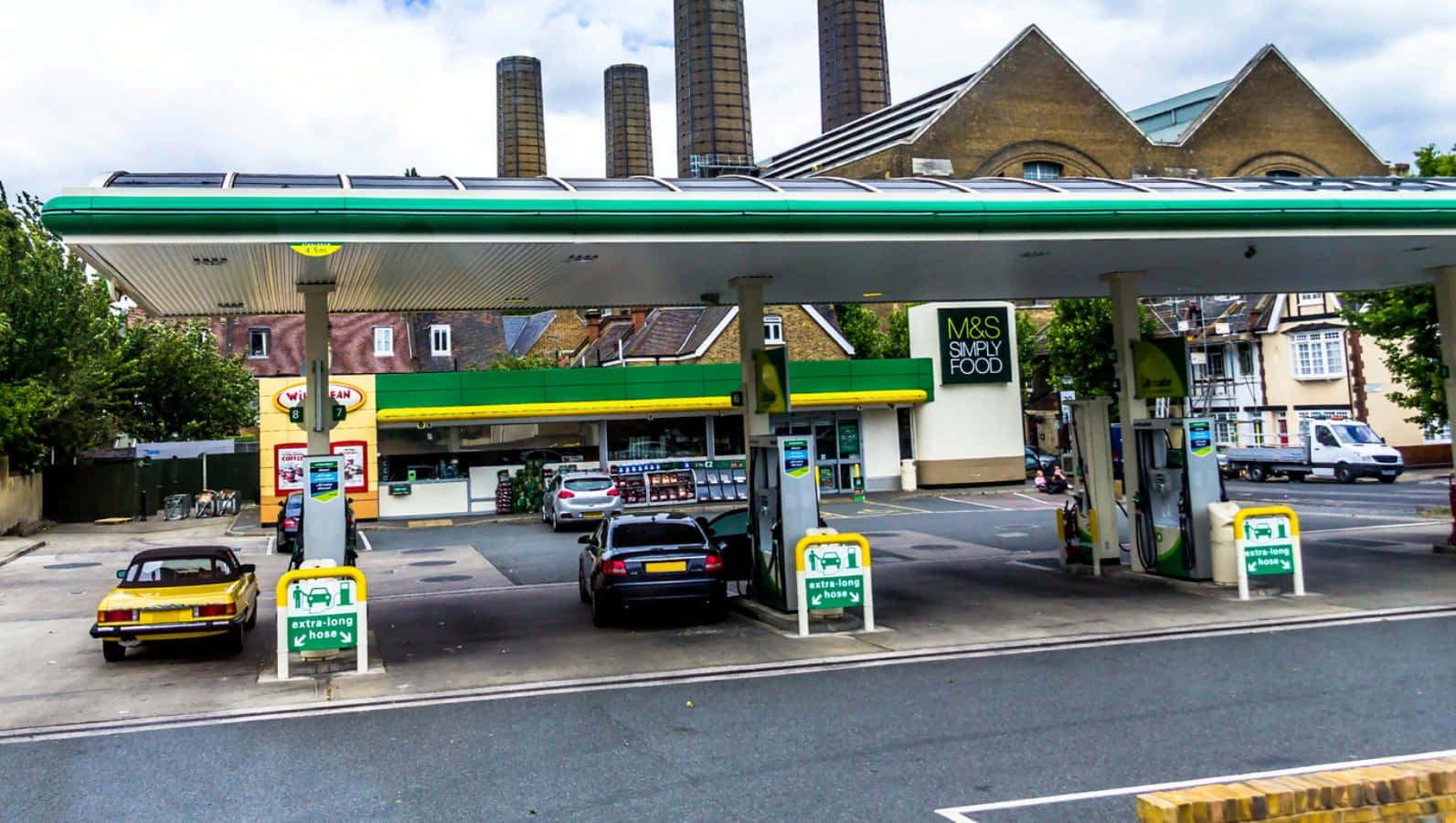 Petrol Station 桌布 壁紙 — 一個綠色和黃色的加油站，有汽車在油泵旁，還有一間M&S Simply Food商店，天空多雲。