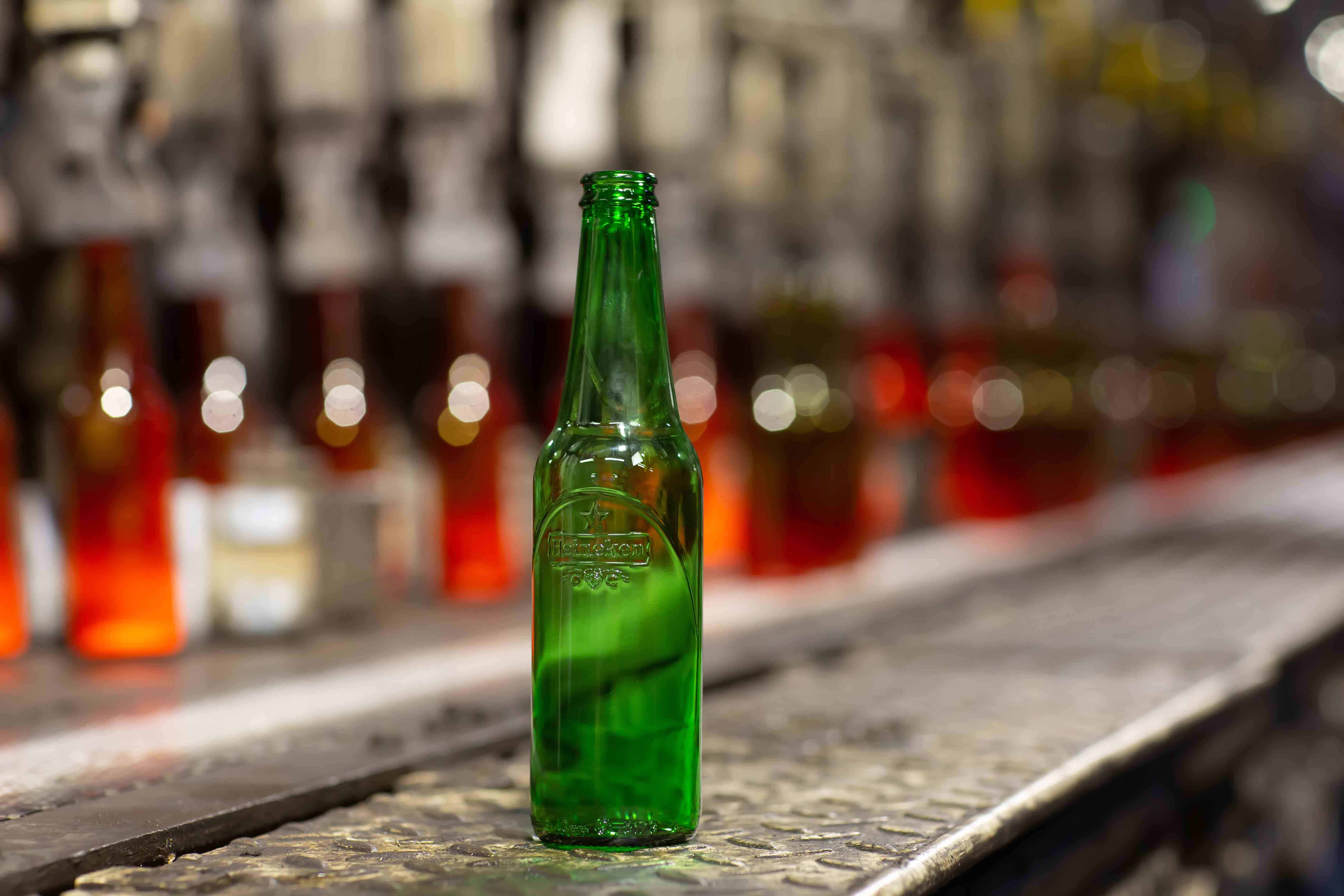 Beer 桌布 壁紙 — 一個綠色啤酒瓶放在有紋理的吧檯上，背景有模糊的瓶子和溫暖的燈光。