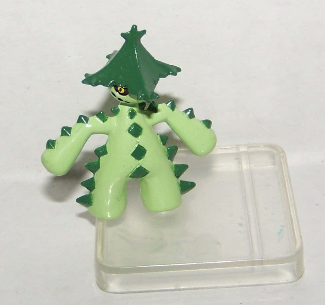 3D 桌布 壁紙 — 一個綠色塑膠公仔，頭部呈樹形，四肢帶有尖刺，站在透明塑膠底座上，背景為純白色。