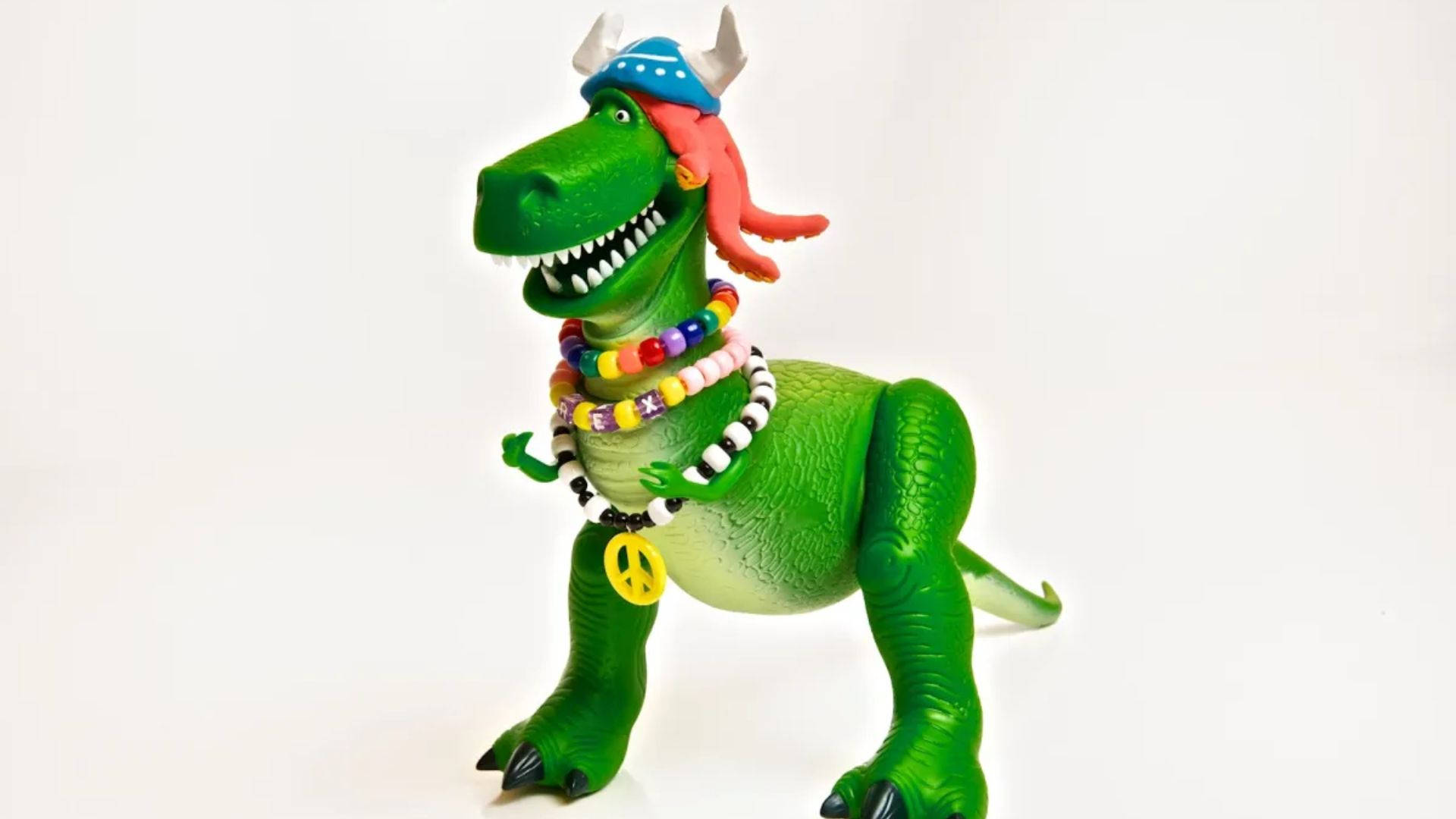 rex 桌布 壁紙 — 一個綠色塑膠恐龍玩具，裝扮成色彩繽紛的嬉皮士，戴著珠子和和平標誌，紅髮，背景為白色。