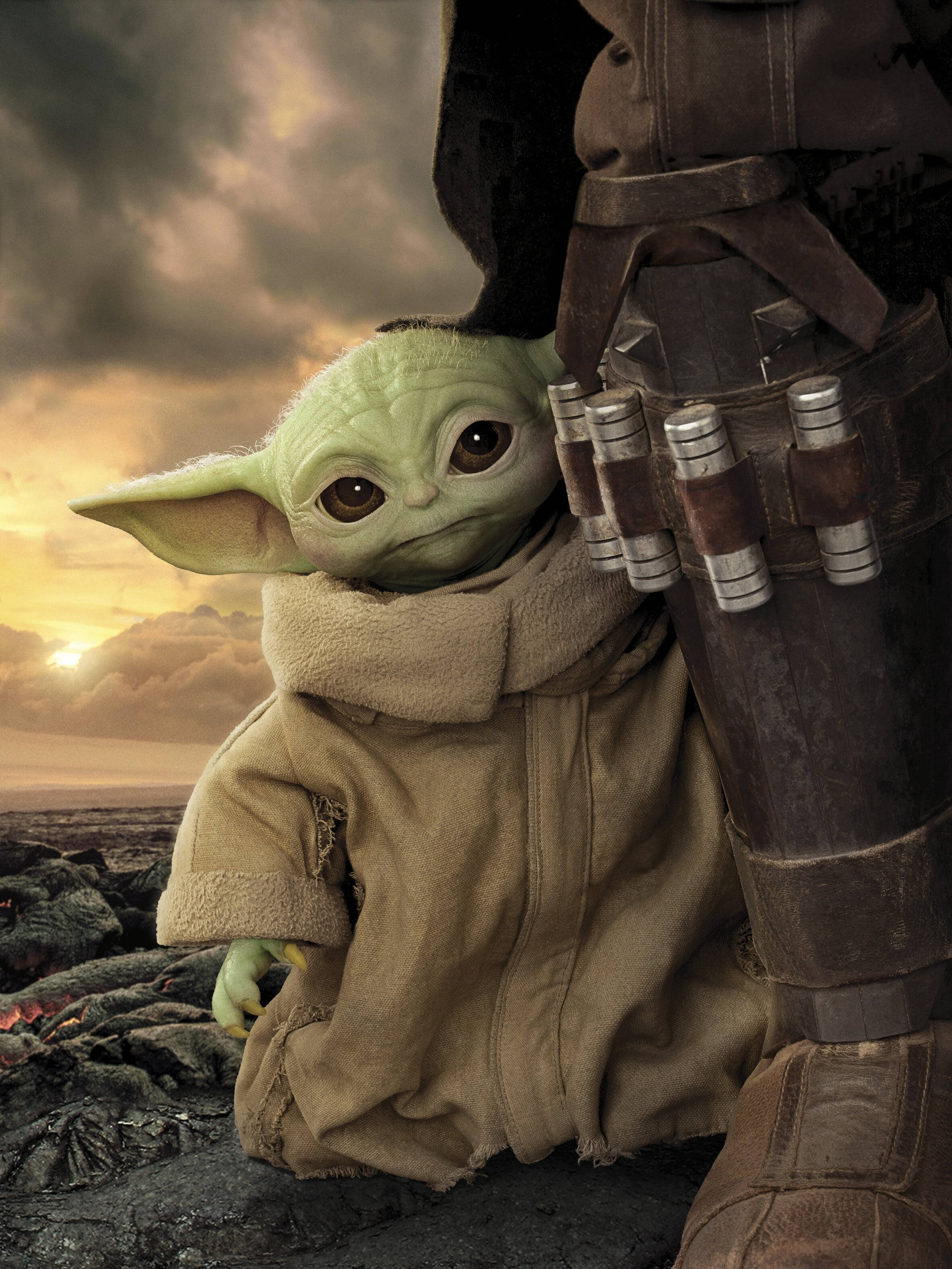 Mandalorian And Baby Yoda 桌布 壁紙 — 一個綠色大眼睛的小孩緊貼著一個裝甲人物，背景是戲劇性的日落天空。