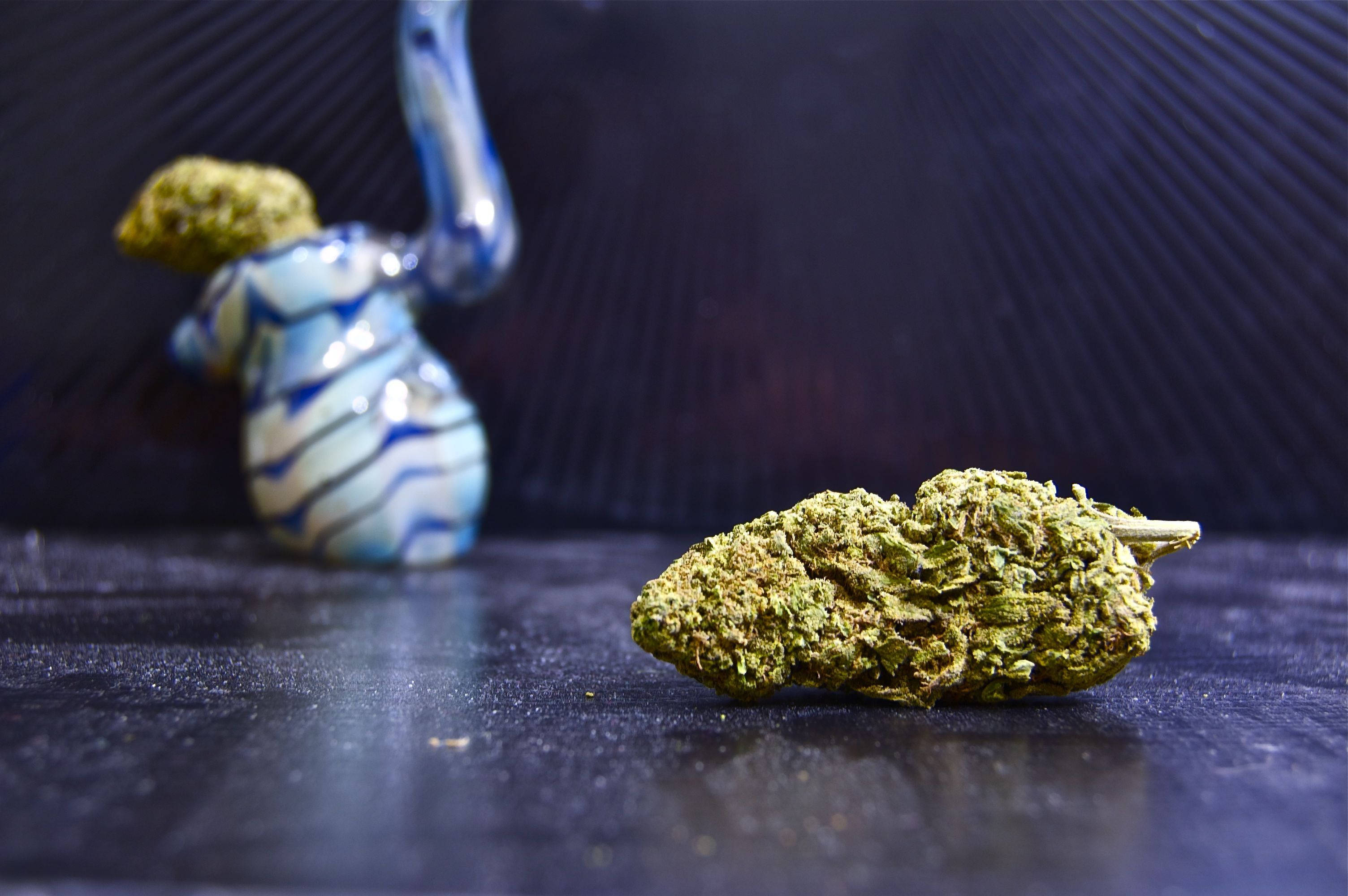 大麻 桌布 壁紙 — 一個綠色大麻花蕾的特寫，放在深色有紋理的表面上，背景模糊中有一個藍色玻璃水煙槍。
