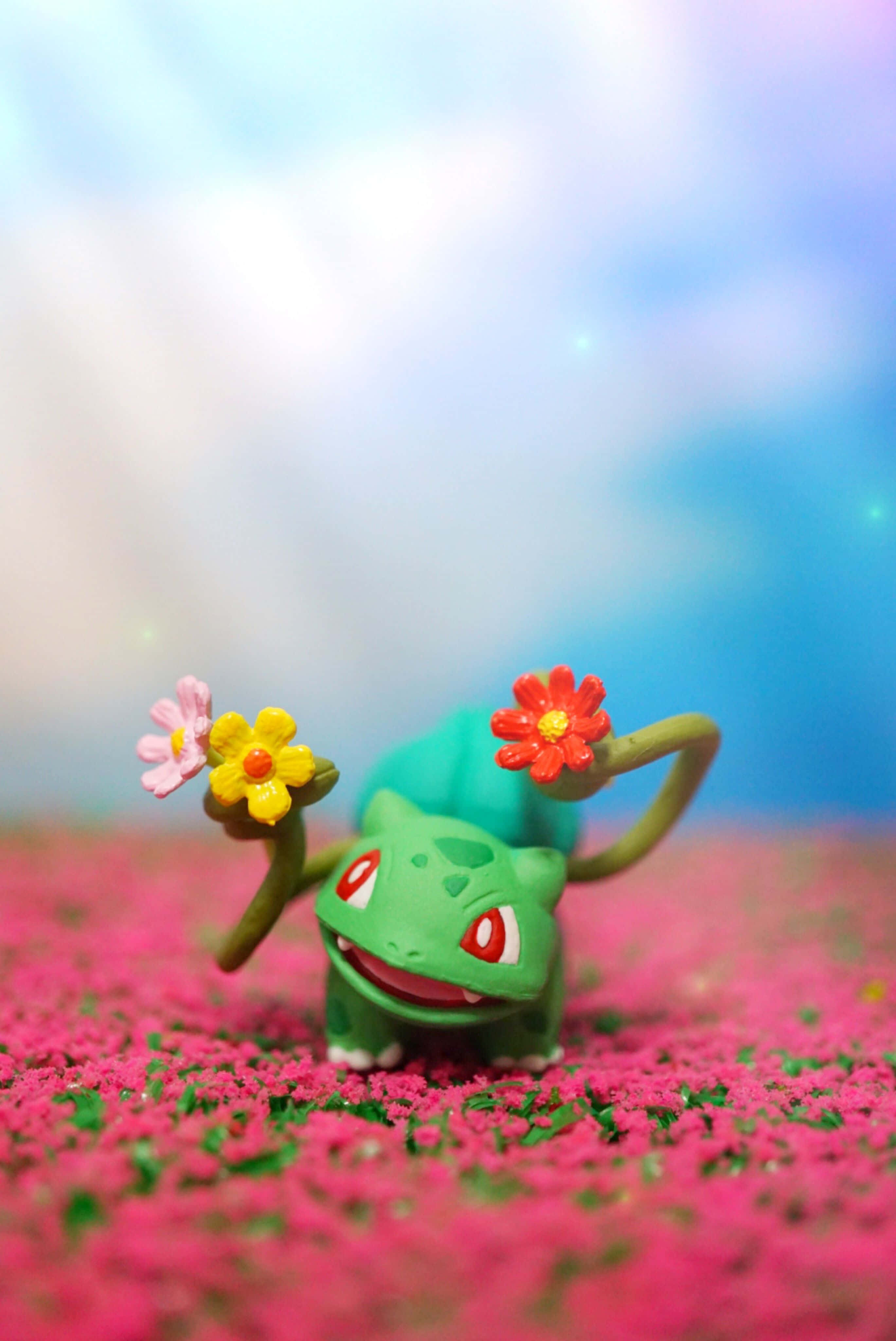 Pokemon Figures 桌布 壁紙 — 一個綠色妙蛙種子玩具站在粉紅色花田中，手持色彩繽紛的花朵，背景是柔和的藍天。