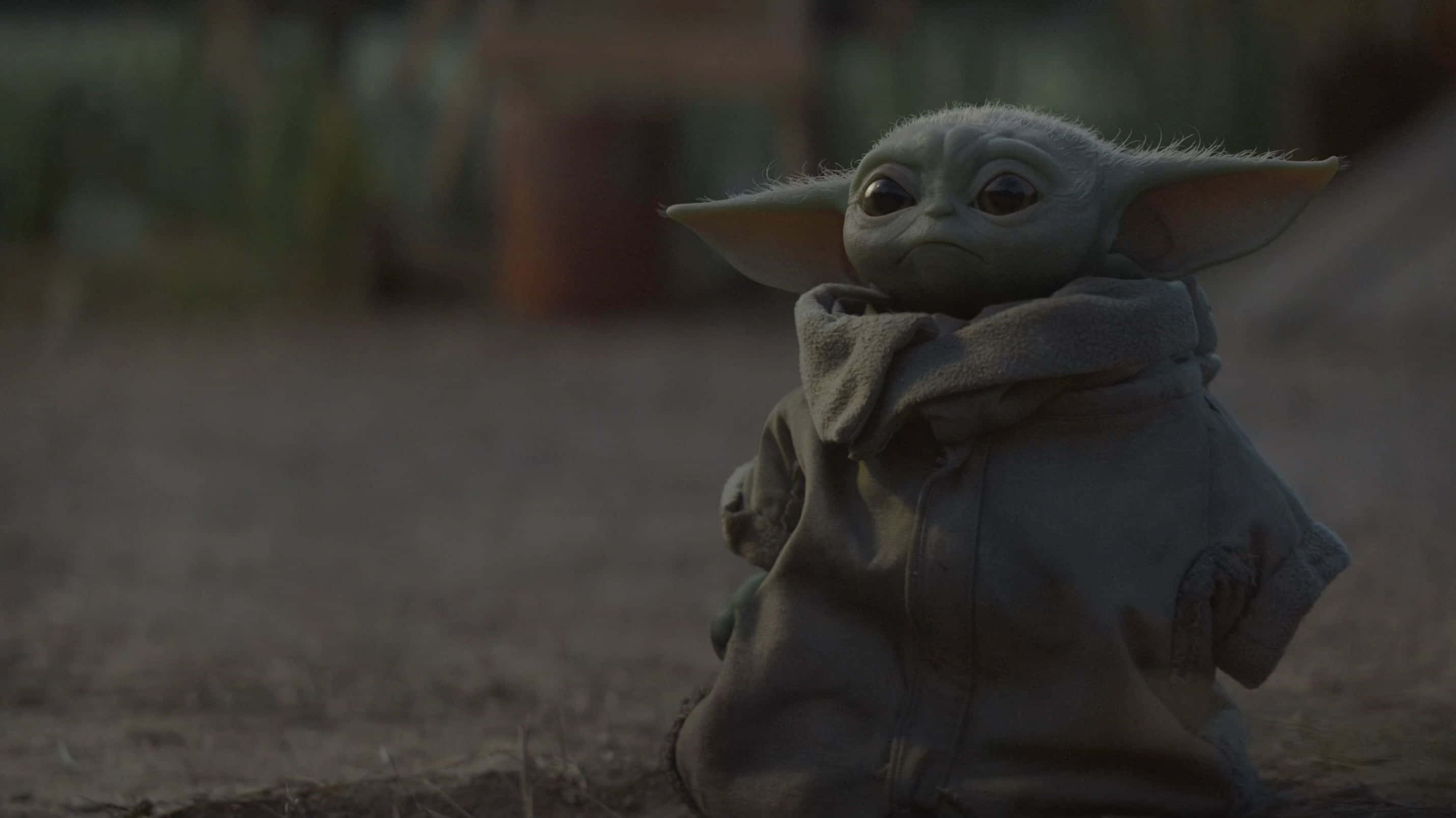 Mandalorian Yoda 桌布 壁紙 — 一個綠色小外星人小孩，耳朵很大，裹著圍巾坐在塵土飛揚的地上，光線昏暗。