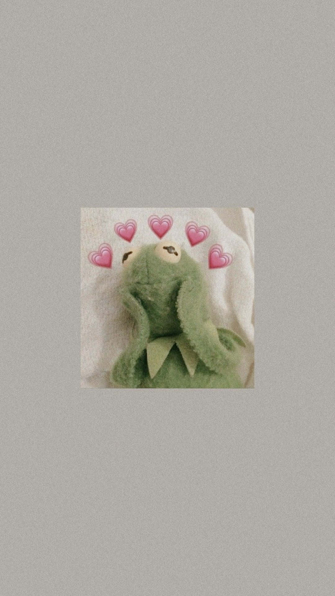 Aesthetic Kermit 桌布 壁紙 — 一個綠色毛絨青蛙玩具，頭頂上方漂浮著粉紅色心形表情符號，背景為白色紋理。