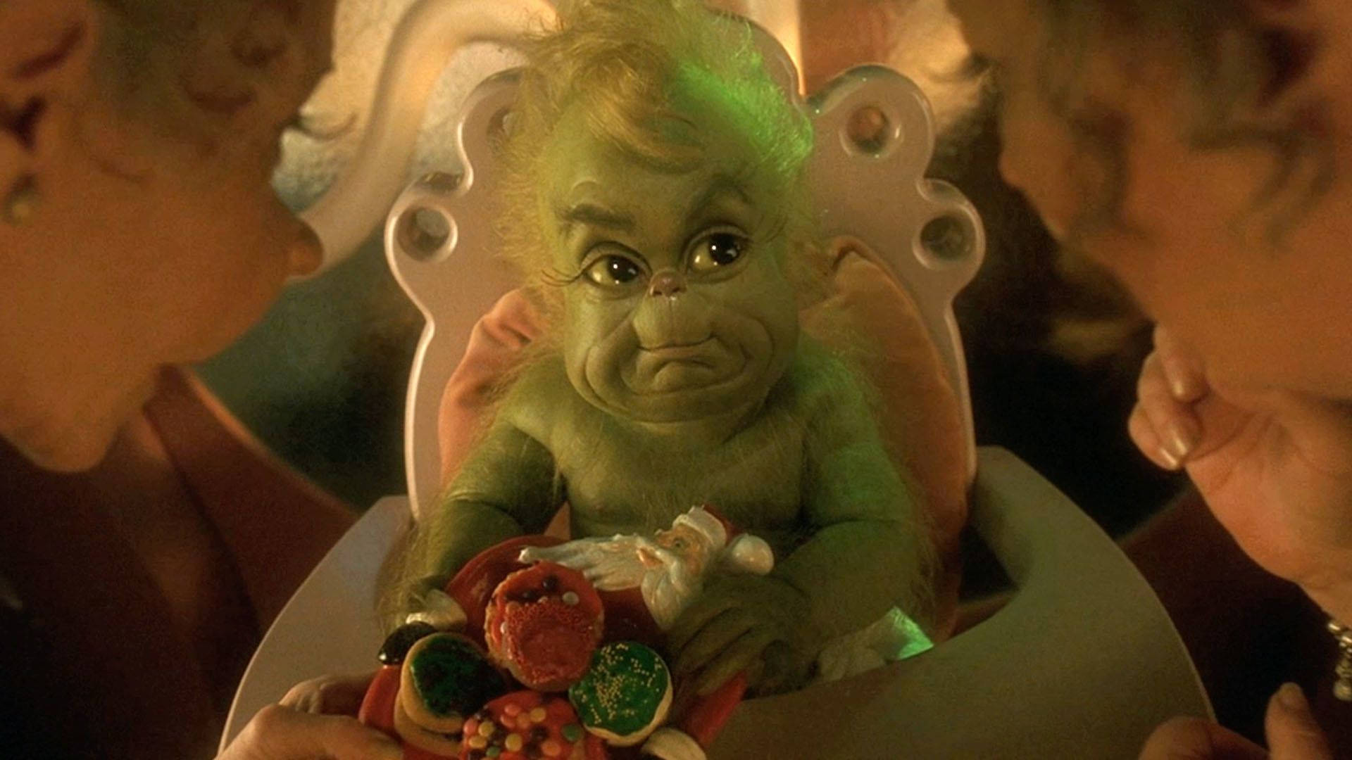 Baby Grinch 桌布 壁紙 — 一個綠色毛茸茸的嬰兒聖誕怪傑坐在高腳椅上，周圍是拿著餅乾的成年人。