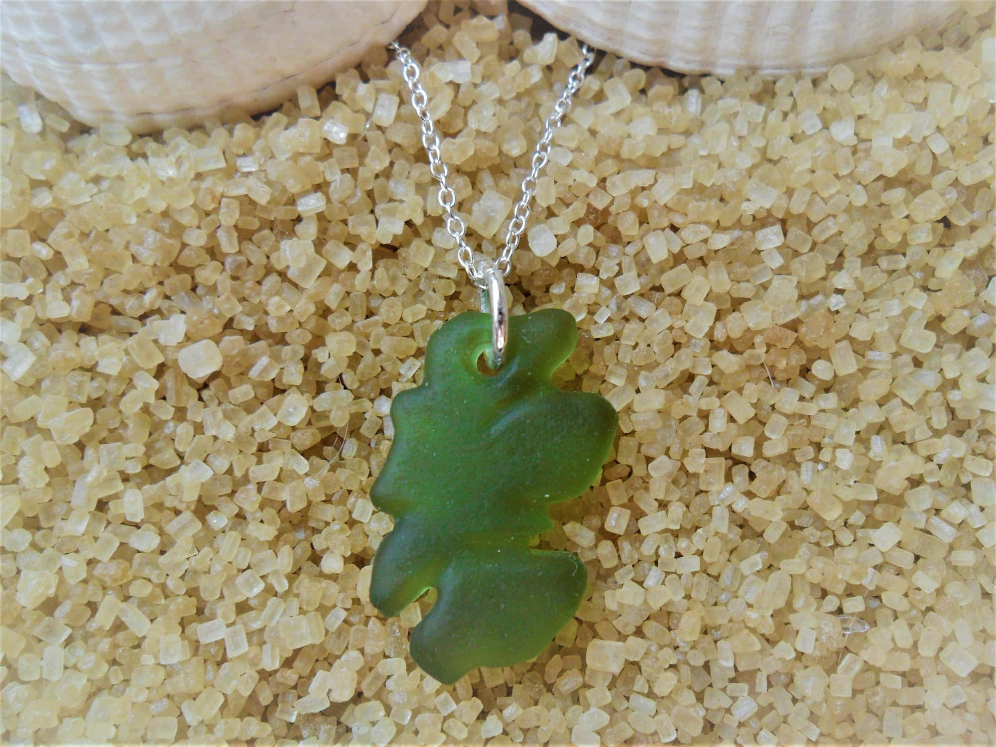 Sea Glass 桌布 壁紙 — 一個綠色海玻璃葉子吊墜放在粗糙的金色沙子上，旁邊有兩個貝殼。