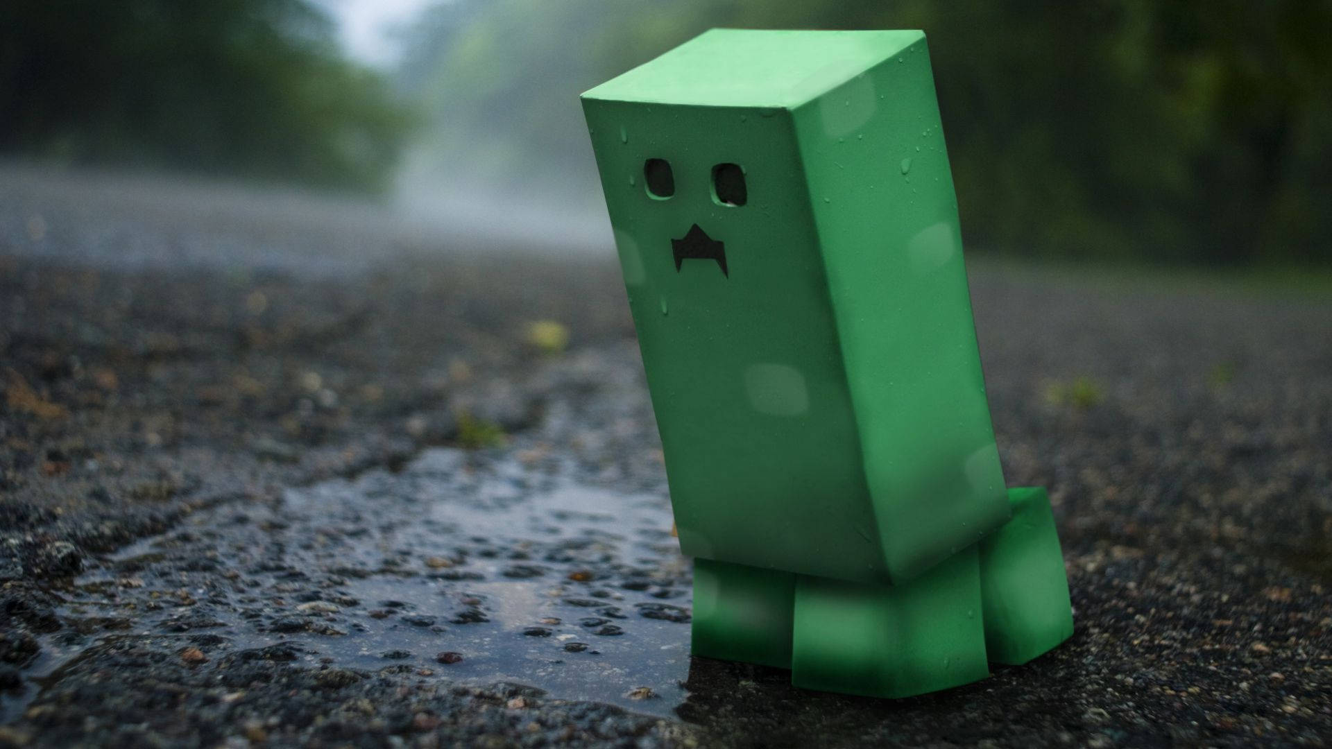 4K Minecraft 壁紙 桌布 壁紙 — 一個綠色爬行者公仔獨自站在潮濕的瀝青路上，背景是模糊的森林，天氣下雨。