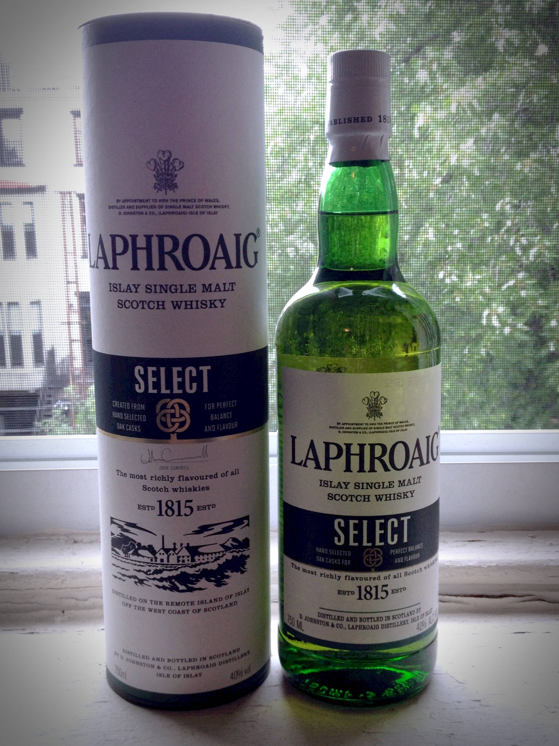 iPhone 桌布 壁紙 — 一個綠色玻璃瓶和圓柱形盒子的Laphroaig Select Islay Single Malt Scotch Whisky並排放在窗台上，後面是模糊的城市景觀。