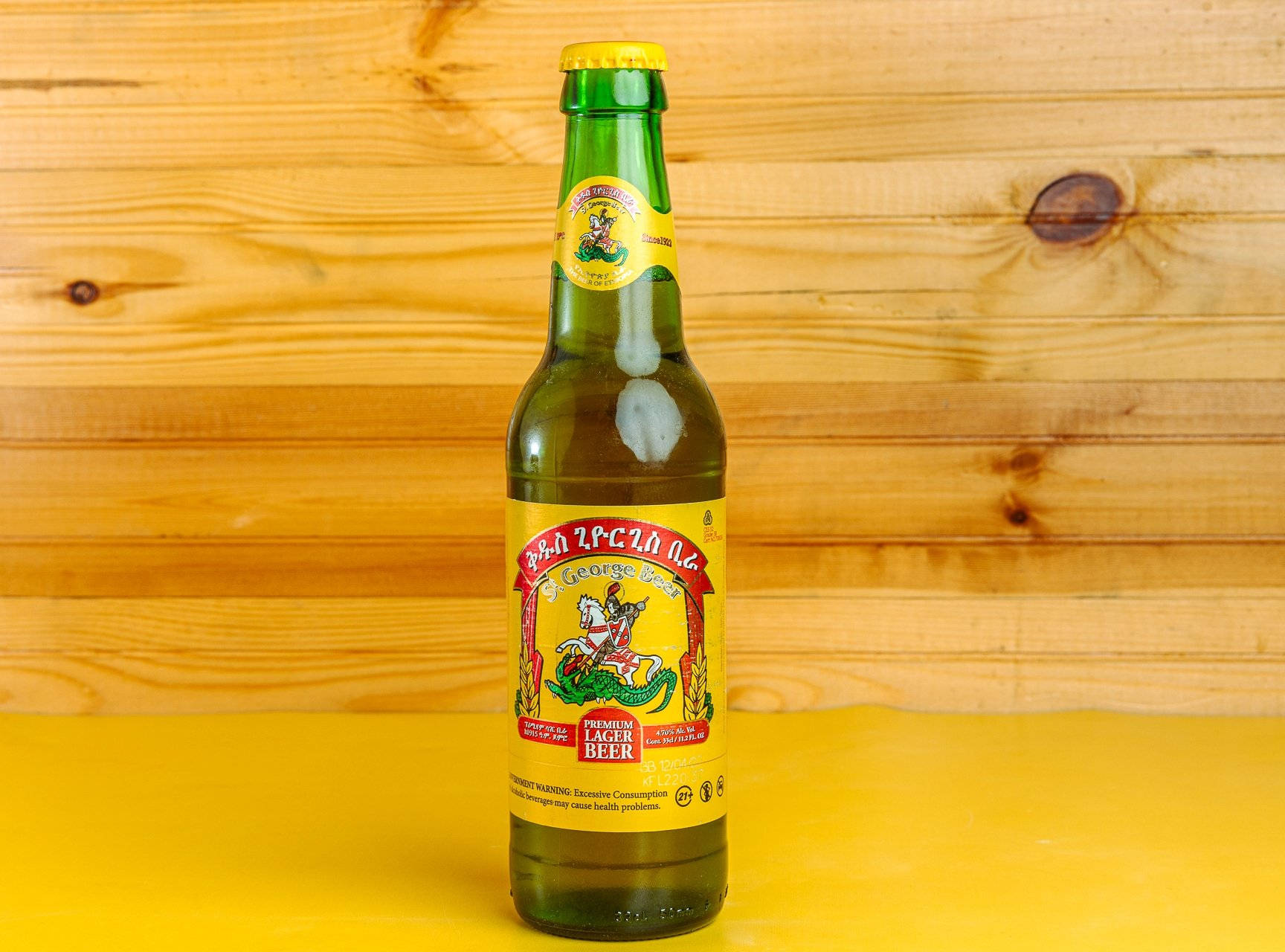 Beer 桌布 壁紙 — 一個綠色玻璃瓶的喬治亞拉格啤酒，黃色標籤上印有騎馬騎士圖案，放在亮黃色表面上，背景為溫暖的木質背景。