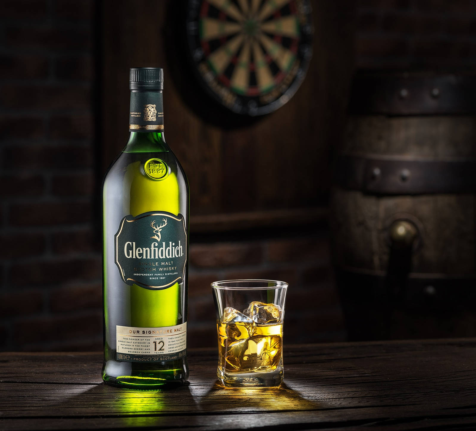 bottle 桌布 壁紙 — 一個綠色的 Glenfiddich 威士忌酒瓶和一個有冰塊的酒杯，放在一張木桌上，旁邊有一個飛鏢靶和一個木桶，在一間燈光昏暗的酒吧裡。