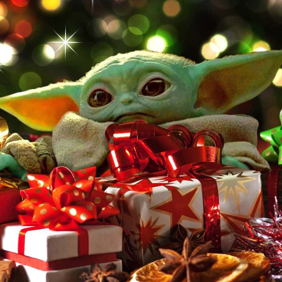 Baby Yoda Christmas 桌布 壁紙 — 一個綠色的Baby Yoda毛絨玩具坐在節日紅色蝴蝶結和星星圖案禮物之間，在閃爍的節日燈光下。