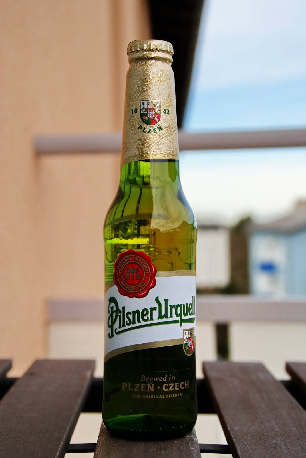 Beer 桌布 壁紙 — 一個綠色的Pilsner Urquell啤酒瓶放在木桌上，背景是模糊的戶外環境。