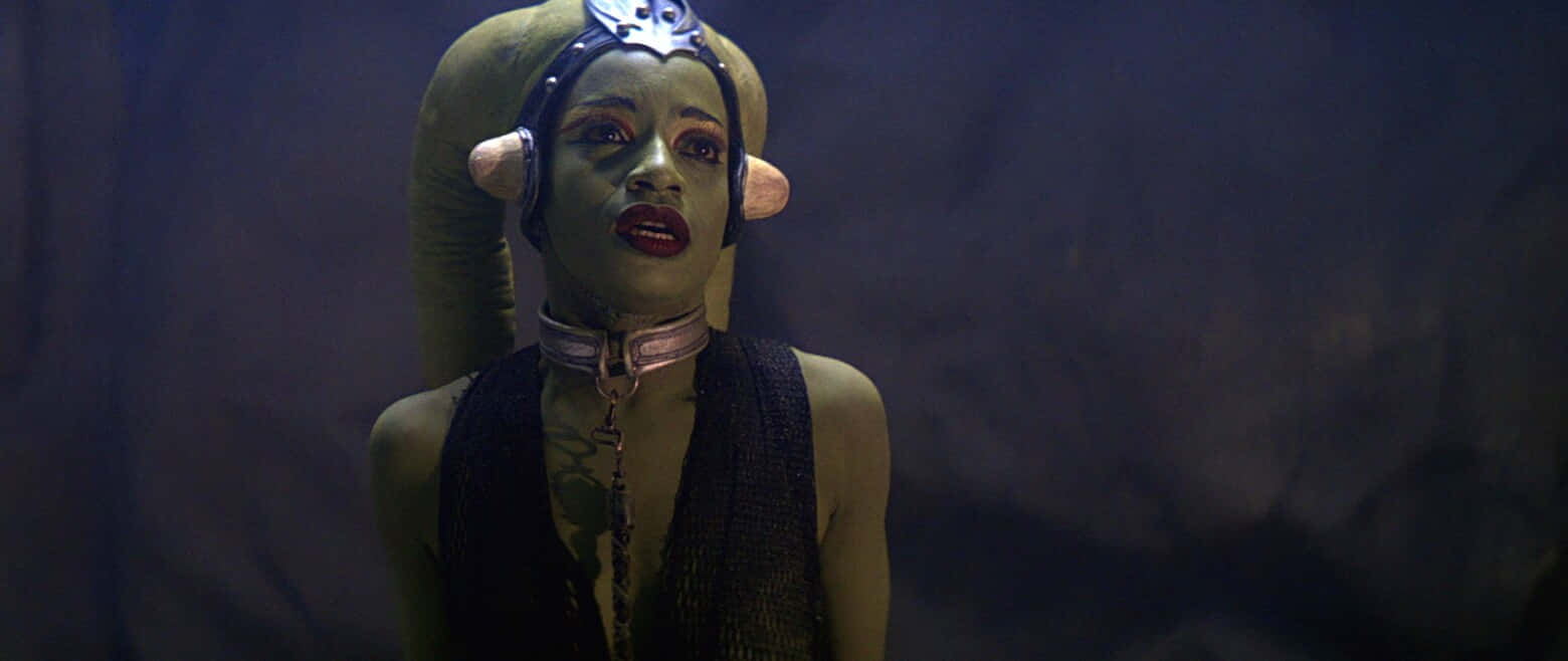 Twi'lek 桌布 壁紙 — 一個綠色皮膚、大耳朵、戴著頭飾的外星女人，在昏暗的洞穴般的環境中專注地凝視。