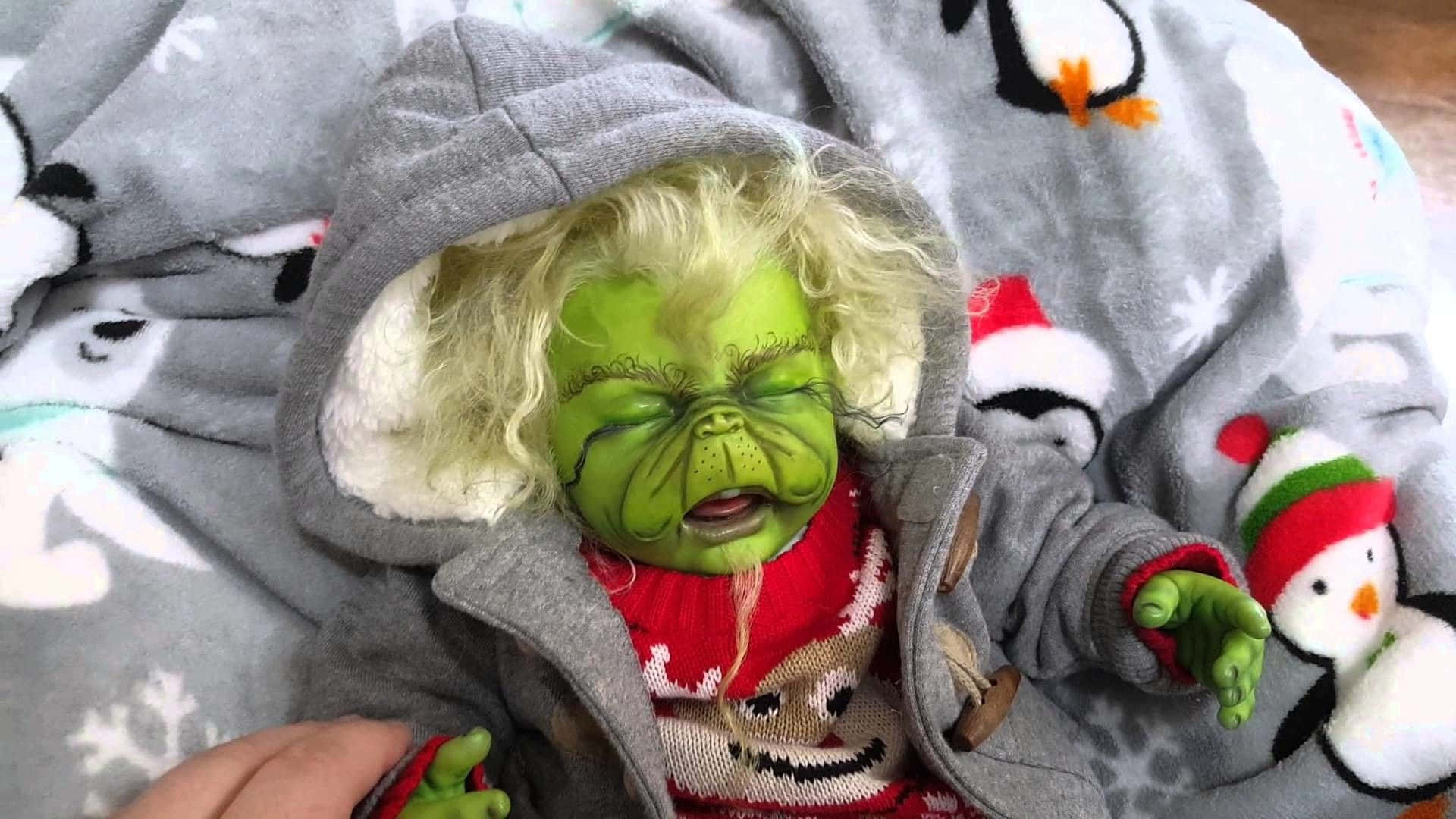The Grinch 桌布 壁紙 — 一個綠色皮膚、金色捲髮的聖誕怪傑公仔，穿著灰色連帽衫和紅色毛衣，在節日毯子上安詳地睡著。