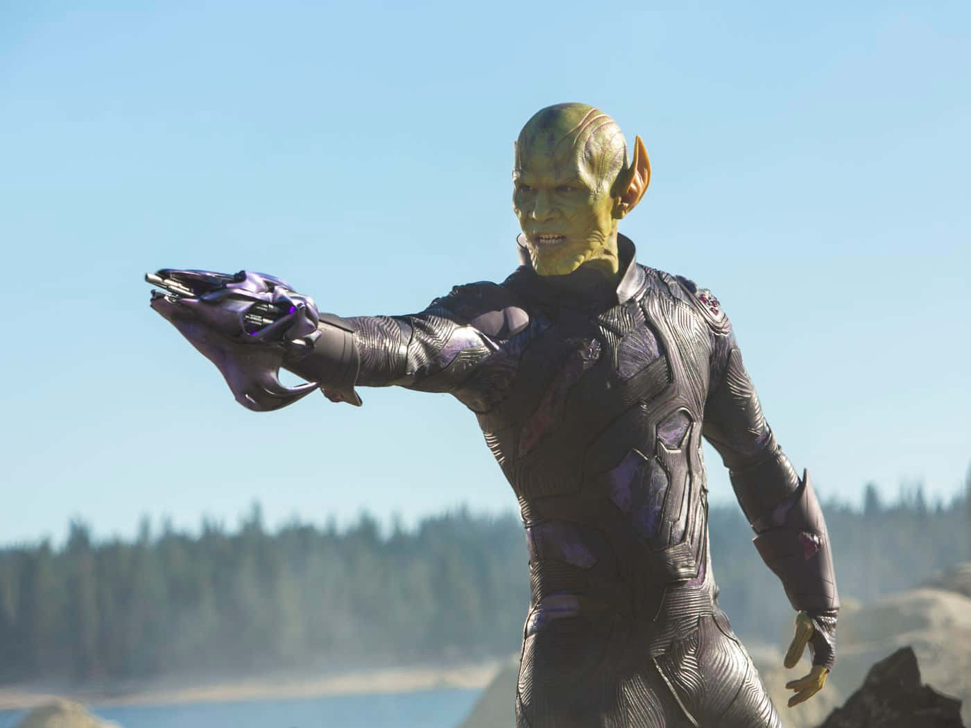 Skrulls 桌布 壁紙 — 一個綠色皮膚的外星戰士穿著時尚的黑色和紫色套裝，在晴朗的藍天下，以森林為背景，伸出發光的武器。