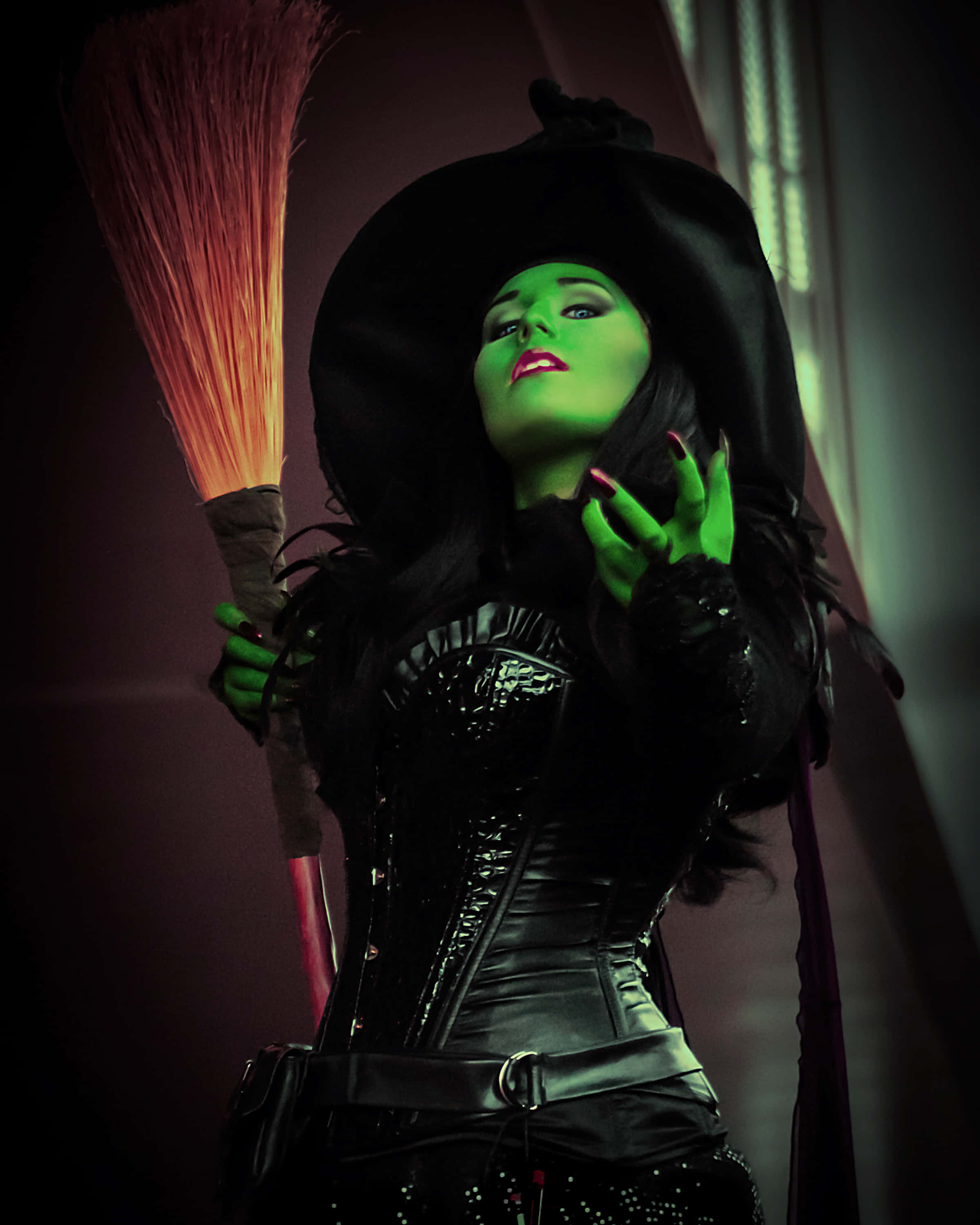 Evil Witch 桌布 壁紙 — 一個綠色皮膚的女巫，戴著黑色帽子和束腰，在昏暗的房間裡舉起發光的手，同時拿著掃帚。