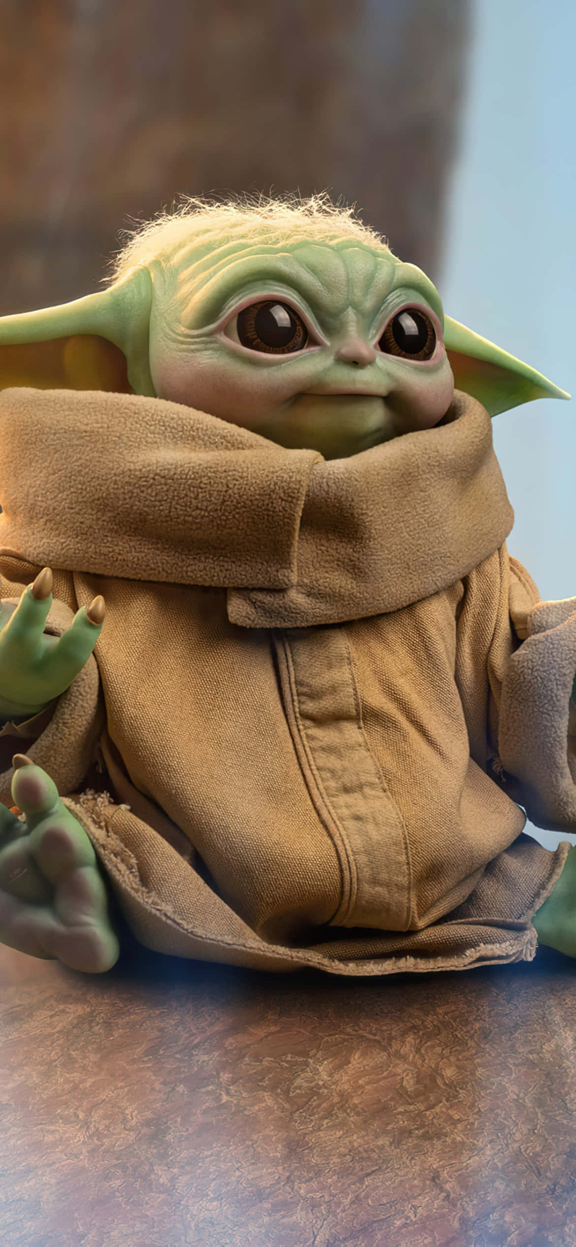 Baby Yoda Phone 桌布 壁紙 — 一個綠色皮膚的嬰兒尤達公仔，包裹著棕色長袍，坐在有紋理的表面上的特寫。